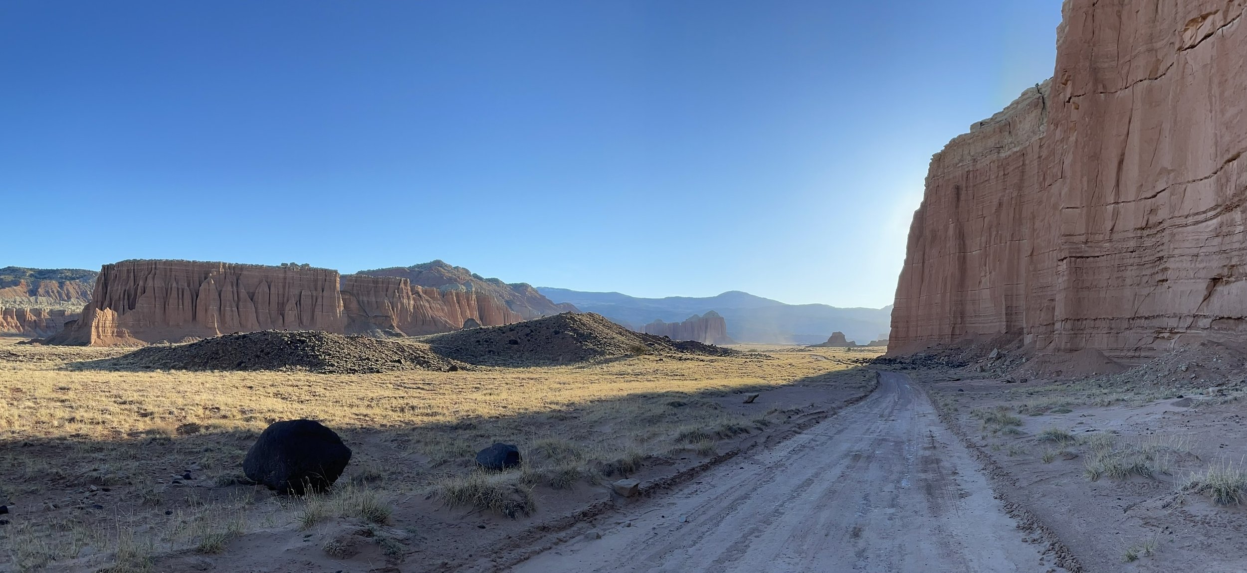 A desert landscape with large red and brown rock formations and cliffs under a clear blue sky. A dirt road runs through the middle of the scene with some scattered rocks and sparse vegetation.