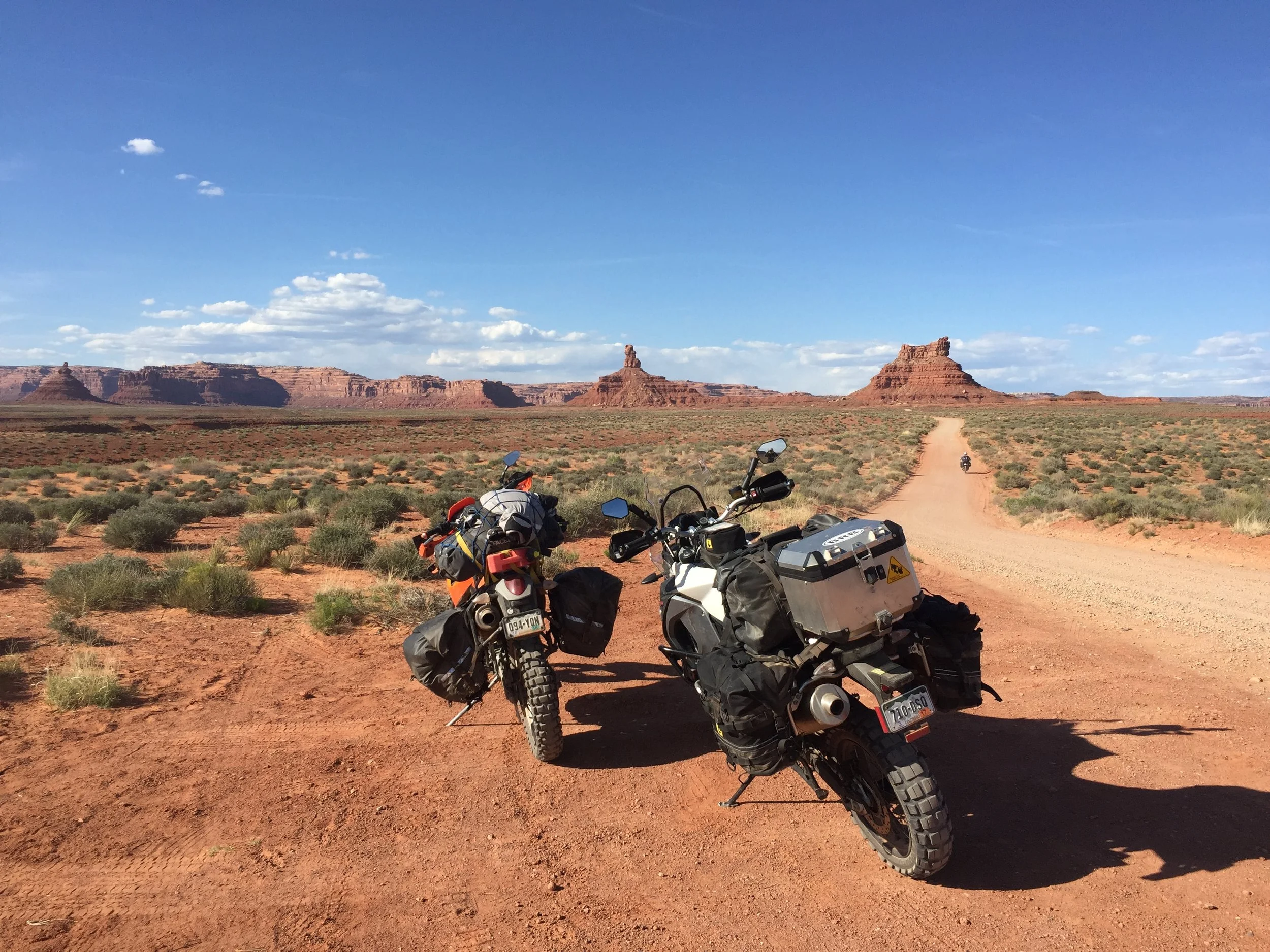 Two adventure motorcycles parked on a dirt trail in a desert landscape with red rock formations in the background and clear blue sky.