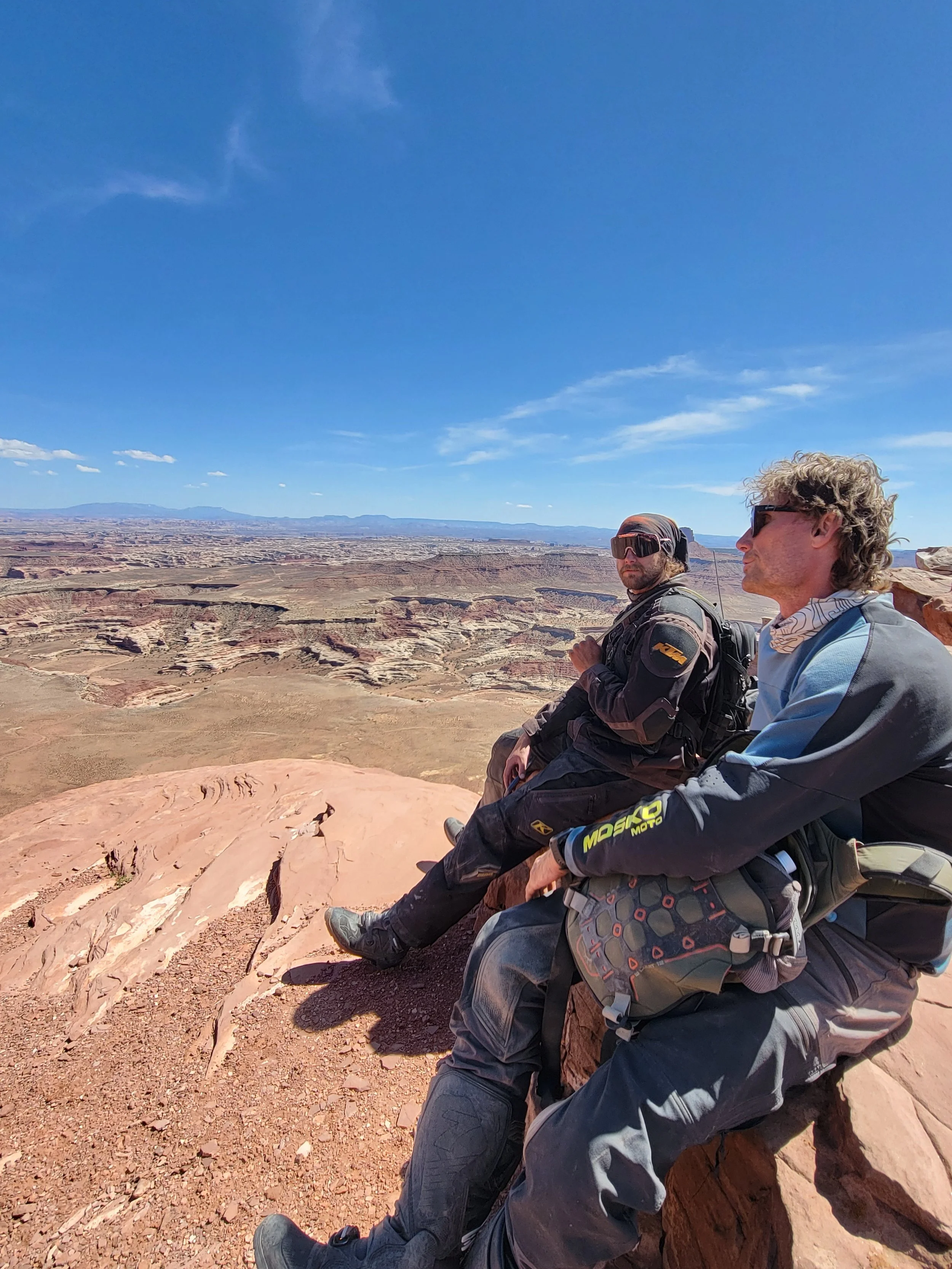 Two men sitting on a rocky ledge overlooking a vast canyon landscape under a clear blue sky.