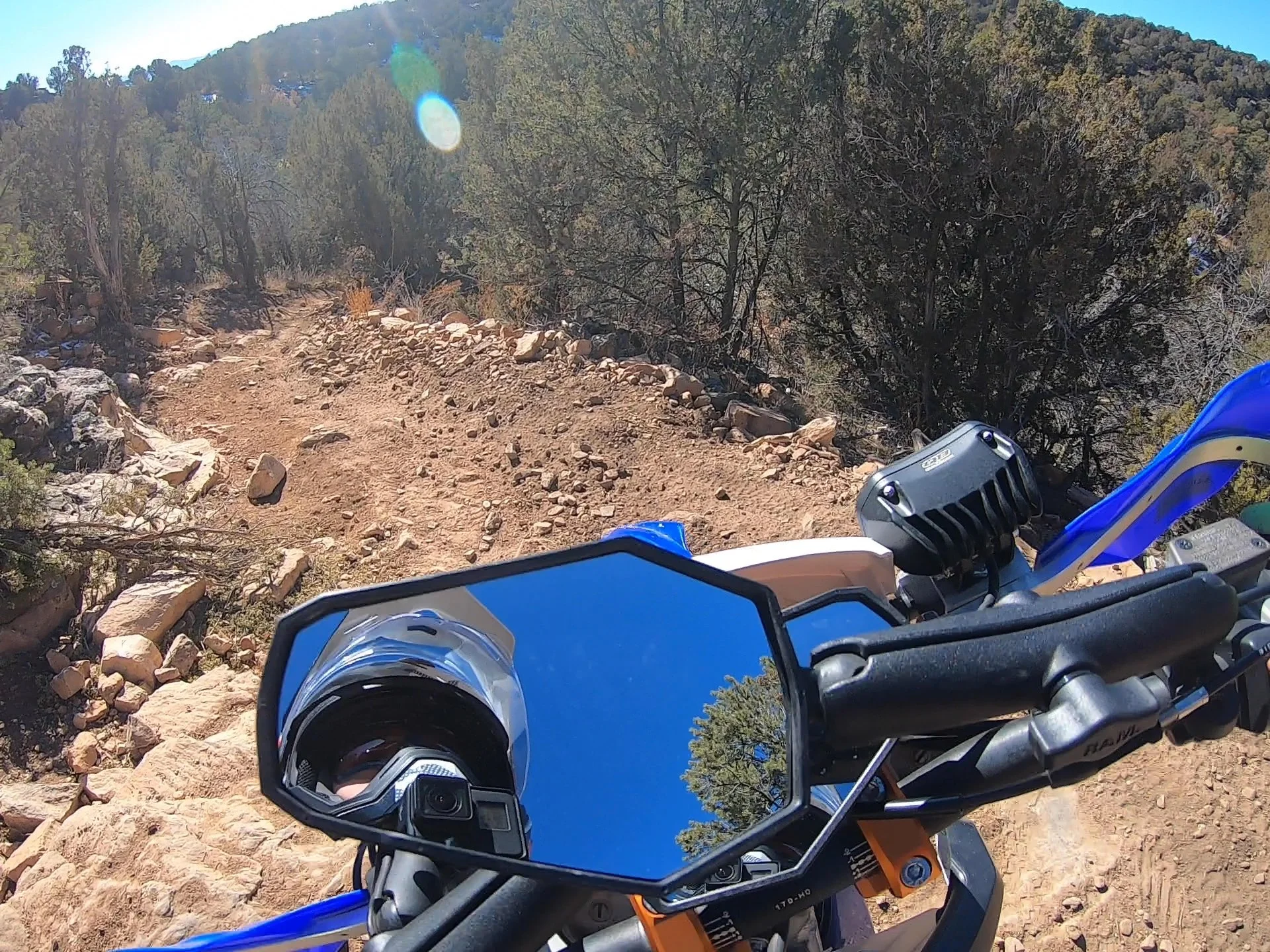 View from a motorcycle rider's perspective on a rocky dirt trail in a forested, mountainous area with trees and hills in the background.