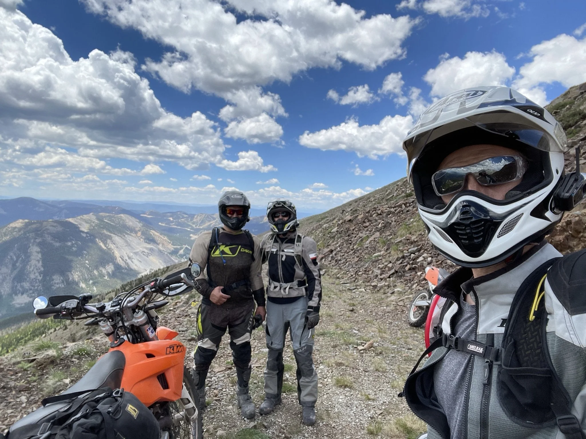Three motorcyclists in gear, standing on a mountain trail with a scenic view of mountains and a partly cloudy sky in the background. One rider is taking a selfie with the other two motorcyclists, all wearing helmets and riding gear, near an orange KTM motorcycle.