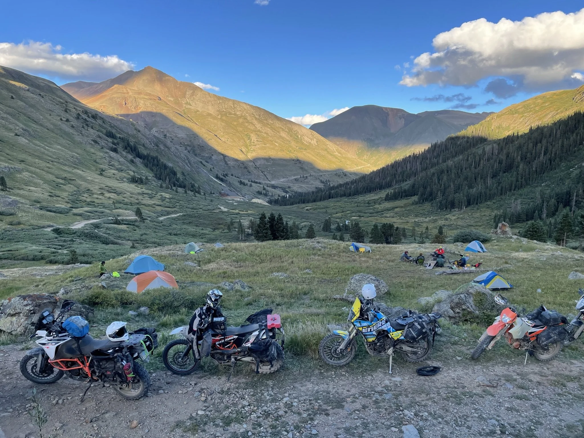 Off-road motorcycles parked on a dirt patch near some tents at a mountain campground, with a scenic valley and mountains in the background under a partly cloudy sky.