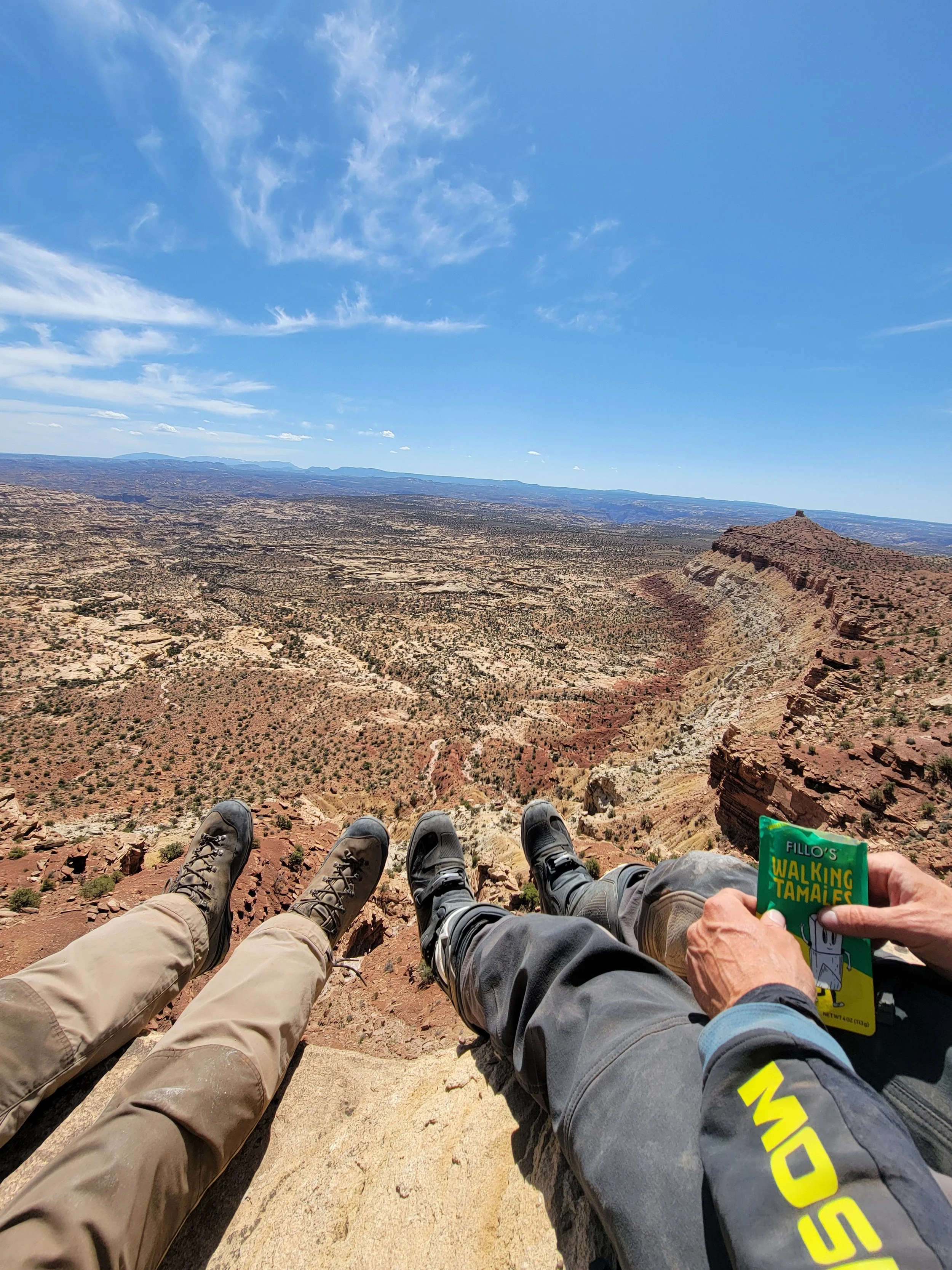 Three people relaxing and enjoying a view of a canyon and desert landscape while sitting on a ledge during a hike, with one person holding a package of tamales.