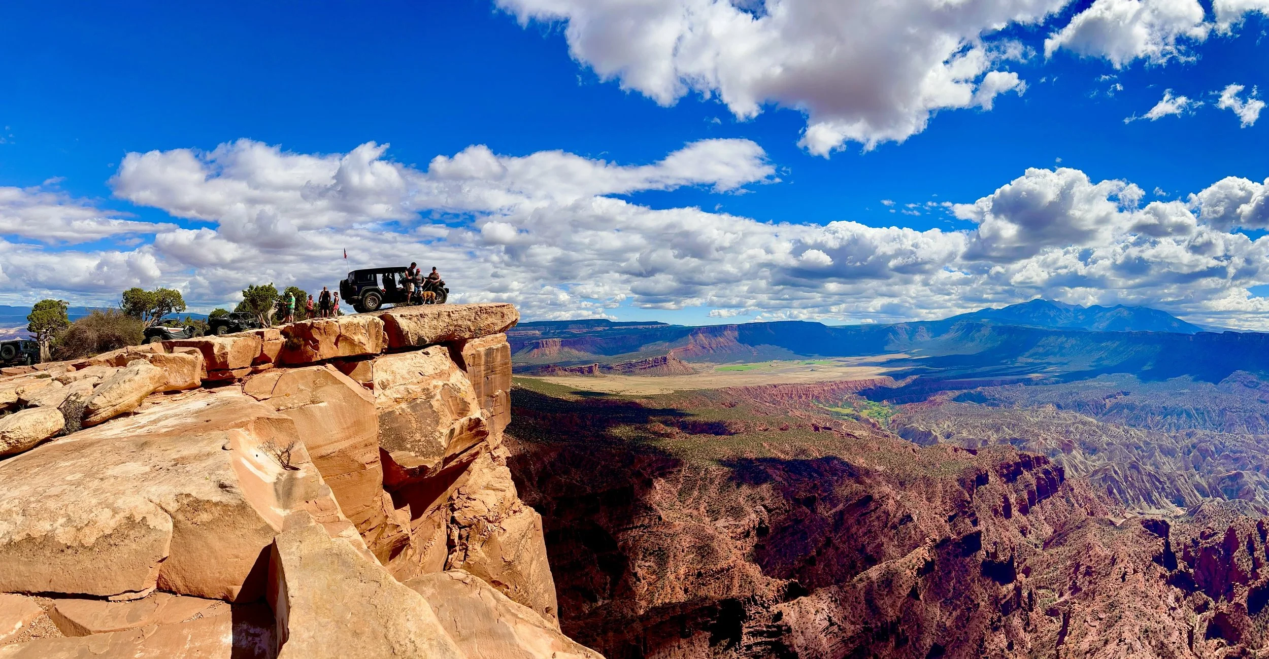 A scenic view of a canyon with red rock cliffs and a vast landscape beyond under a partly cloudy blue sky, with a group of people and vehicles on the edge of a cliff.