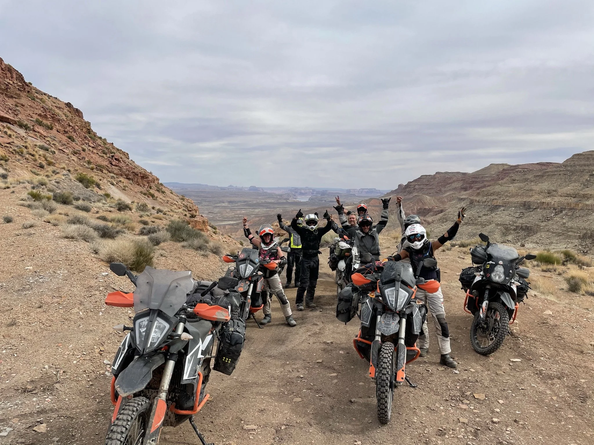 Group of seven motorcyclists wearing helmets and protective gear, standing with their bikes on a dirt trail in a desert canyon landscape, raising their hands in celebration.