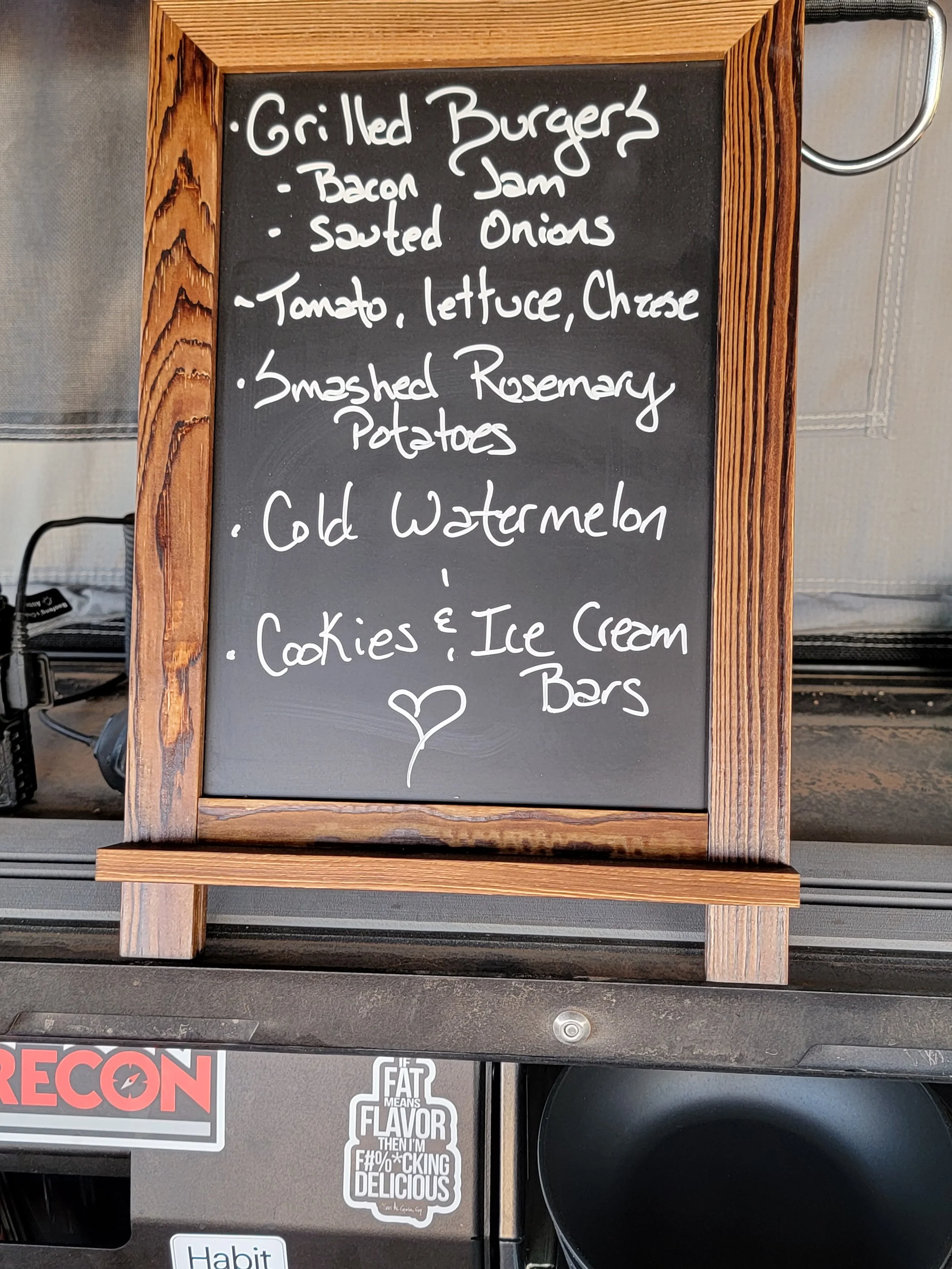 Menu on a black chalkboard with a wooden frame lists food items including grilled burgers with bacon jam, sautéed onions, tomato, lettuce, cheese, smashed rosemary potatoes, cold watermelon, and cookies and ice cream bars, with a small heart drawn at the bottom.