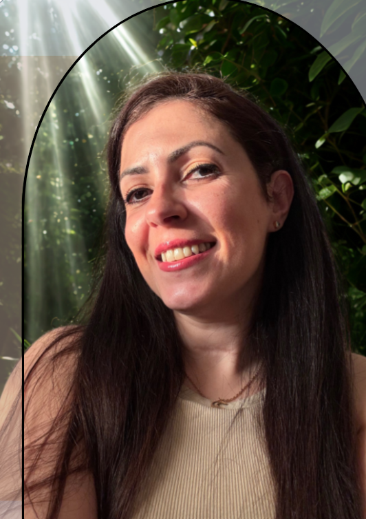 Portrait d'une femme souriante avec de longs cheveux noirs, portant un haut beige, sur fond de forêt avec des rayons de soleil traversant les arbres.