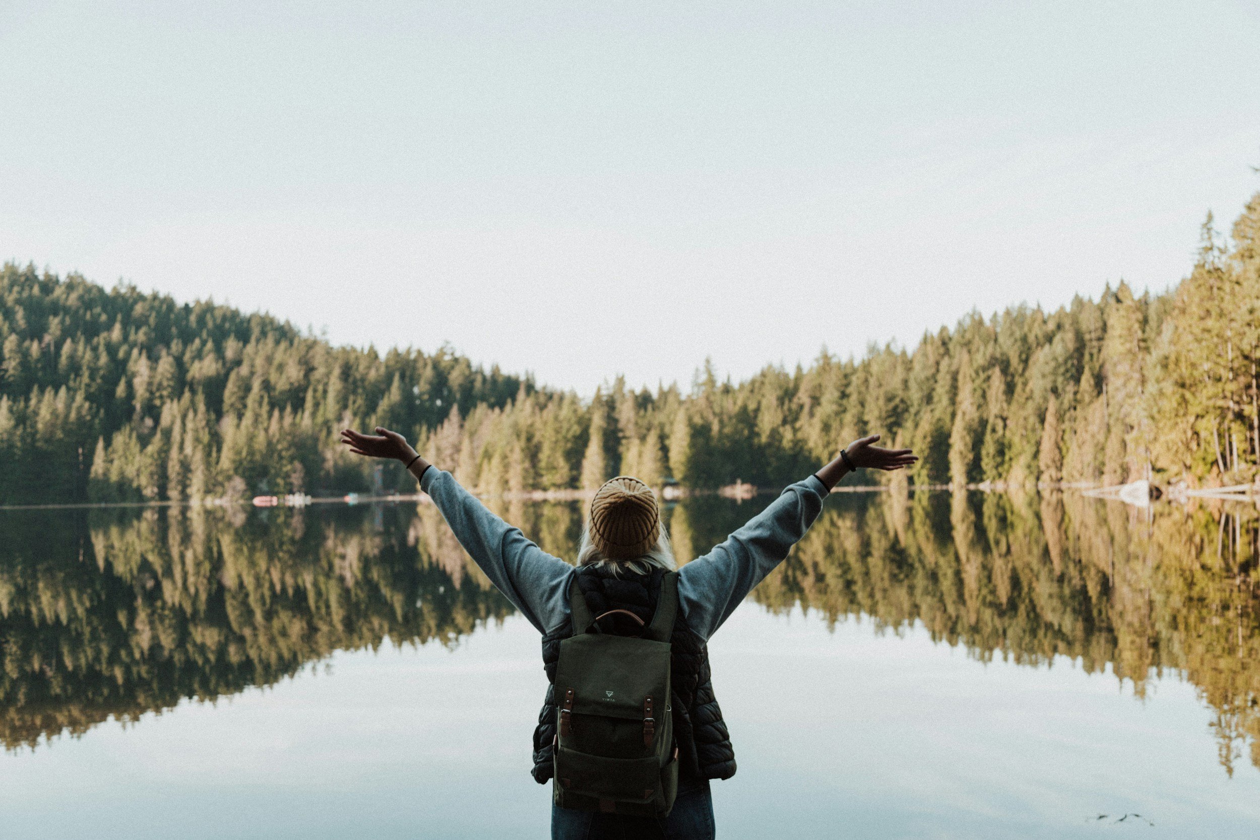 Person with arms raised standing by a calm lake surrounded by forested hills under a clear sky.