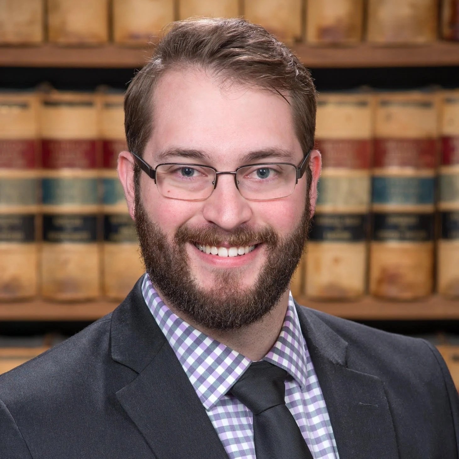 Portrait of a man with glasses, a beard, wearing a suit and tie, in front of a background of shelves filled with law books.