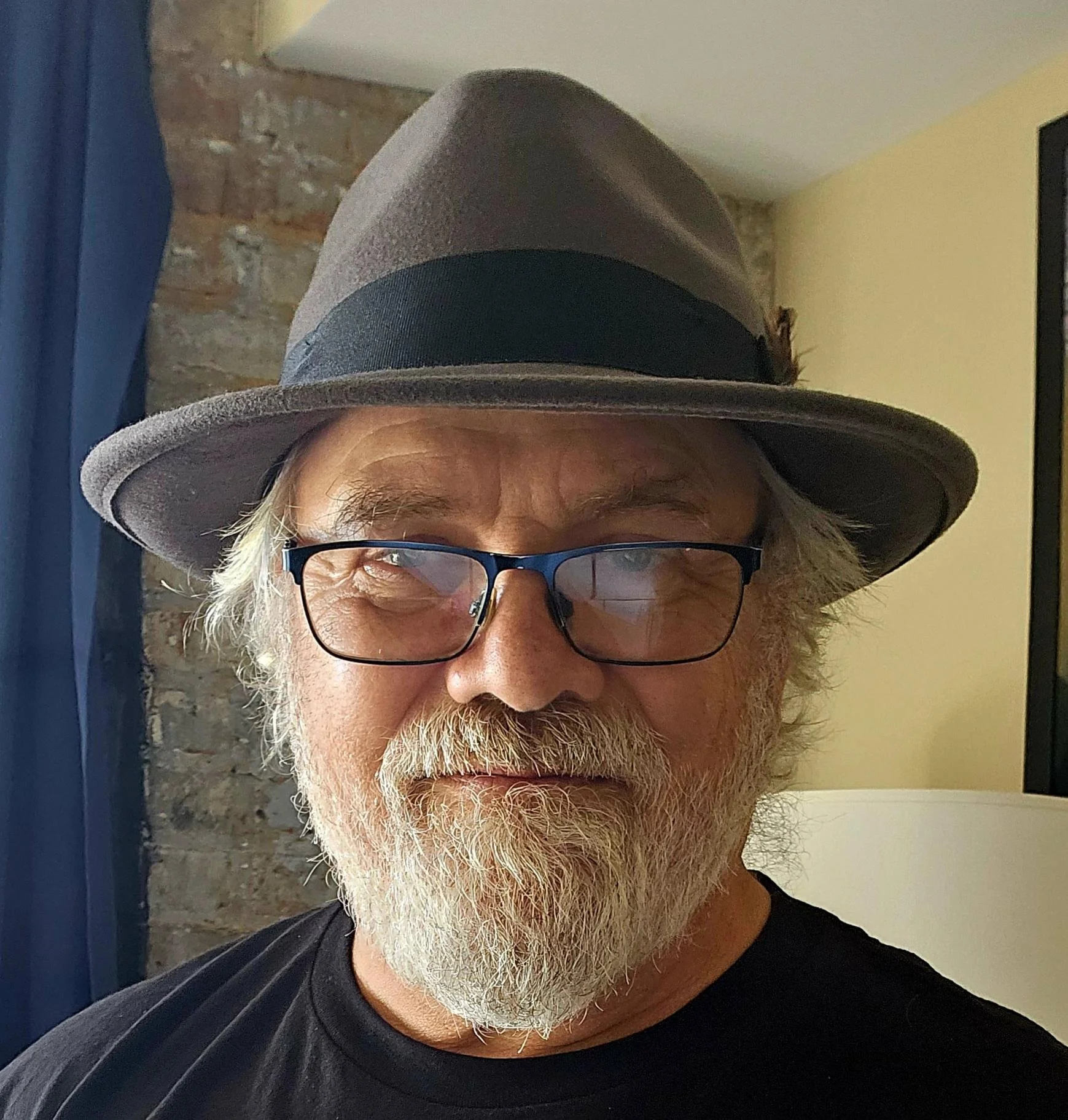 A man with white hair, a beard, and glasses wearing a gray fedora hat with a black band, standing indoors near a brick wall and blue curtains.