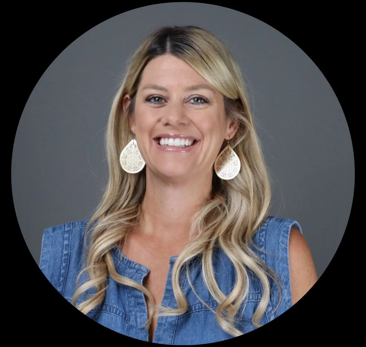 A woman with long blonde hair smiling, wearing large white earrings and a sleeveless denim shirt against a gray background.