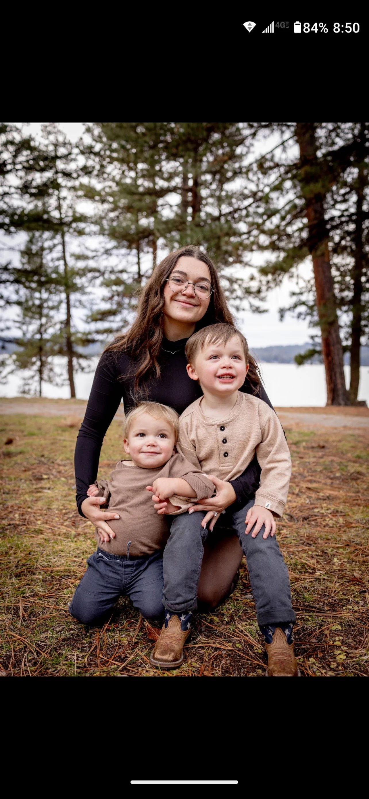 A young woman with glasses and long brown hair kneeling outdoors with two young children, a boy and a girl, in front of a wooded area near a body of water, all smiling.