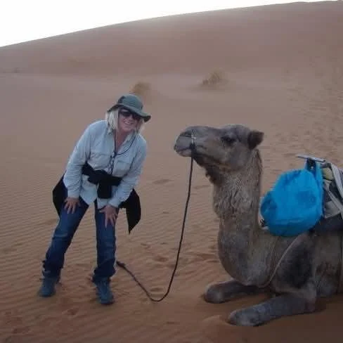 A woman in outdoor clothing and a wide-brimmed hat standing next to a resting camel in a desert with sand dunes in the background.