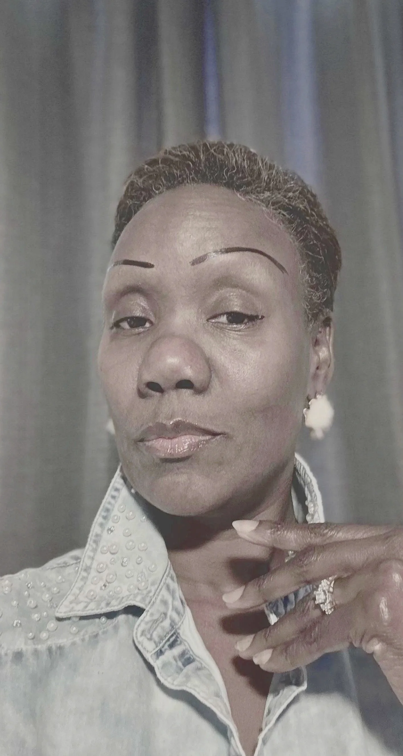 A close-up portrait of a woman with short, styled hair, wearing a light-colored denim shirt with embellishments, a ring, and earrings, posing with her hand near her face against a plain background.