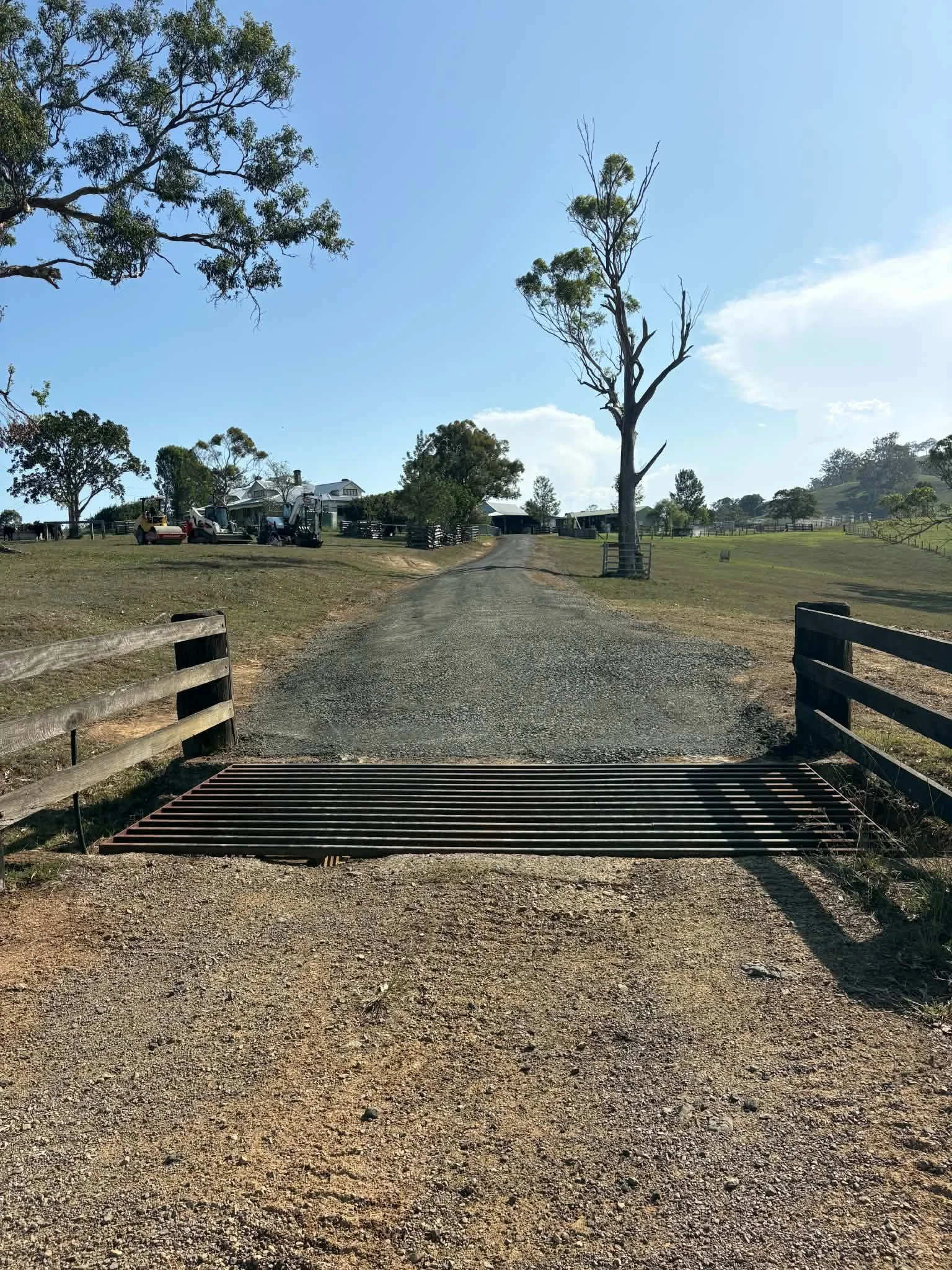 A rural driveway with a metal drainage grate in the foreground, lined with wooden railings, leading past a tree and farm buildings in the background under a clear blue sky.