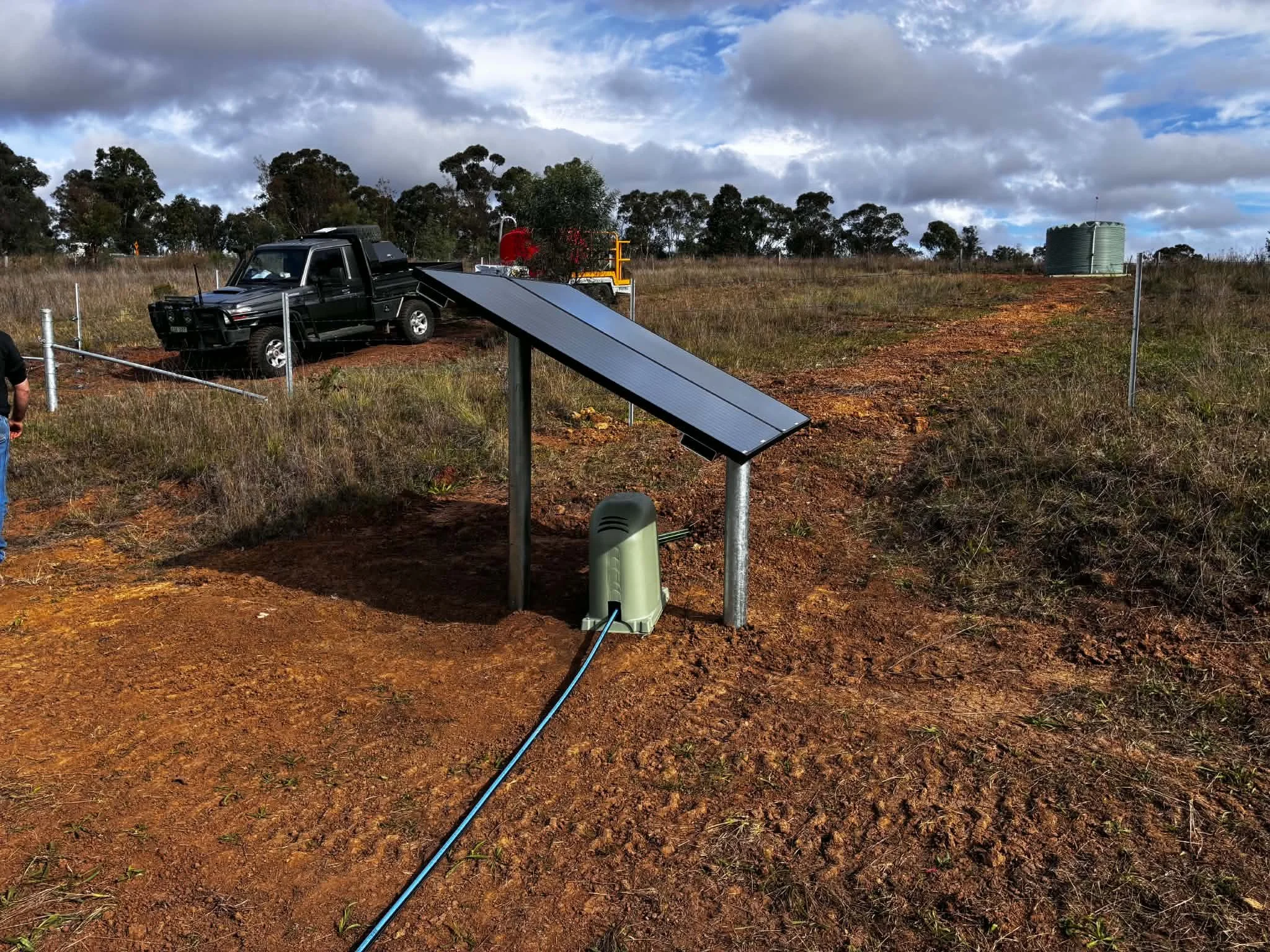 A black utility vehicle parked on a dirt patch with a solar panel next to it. There is a solar water heater or solar thermal collector along the ground connected to a power source, with a solar water tank visible on the right. The setting is a rural 