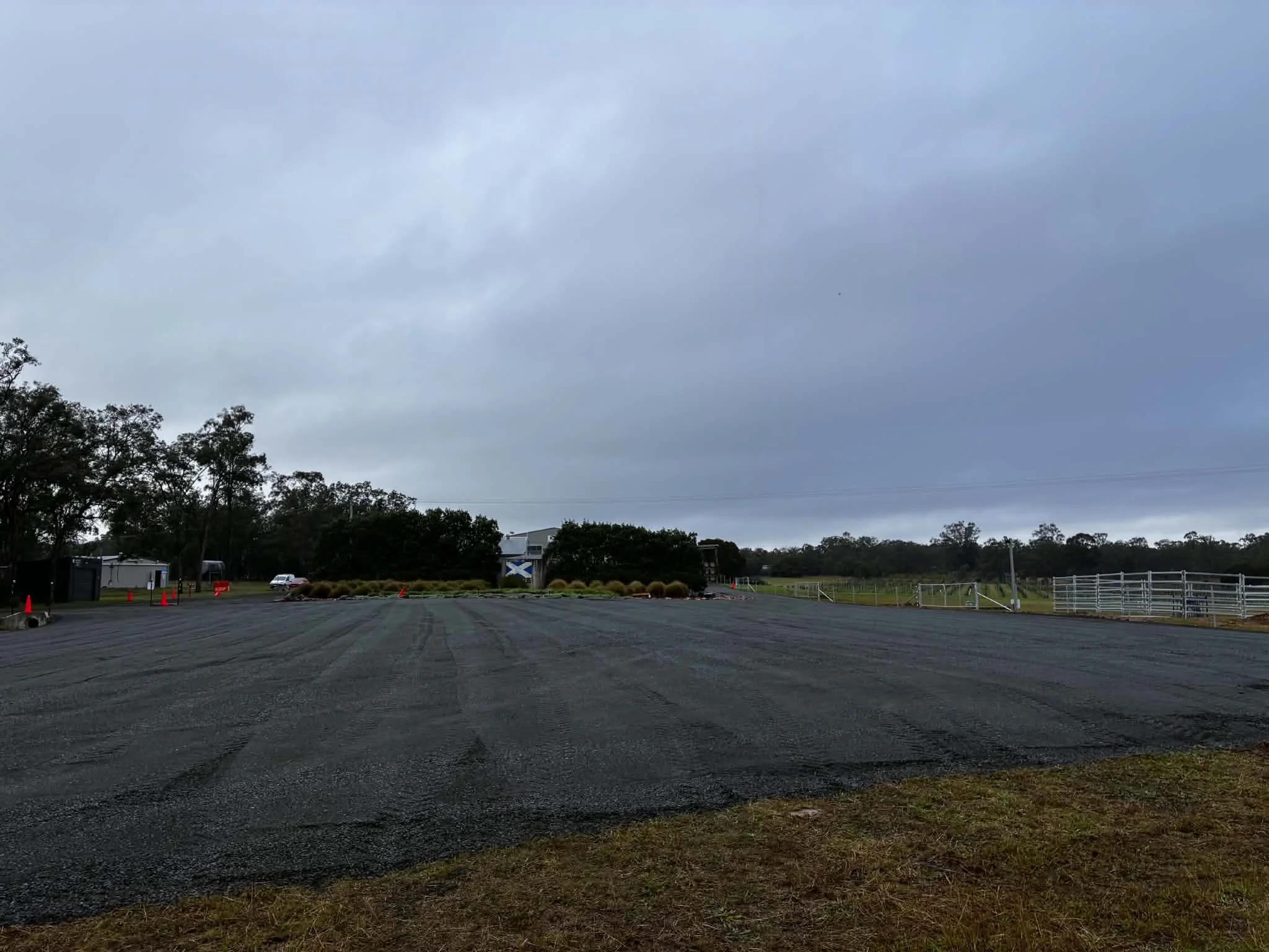 An outdoor parking lot with a cloudy, overcast sky. The lot is mostly empty, with a few cones and some vehicles. There are trees and a building with a blue and white sign in the background.