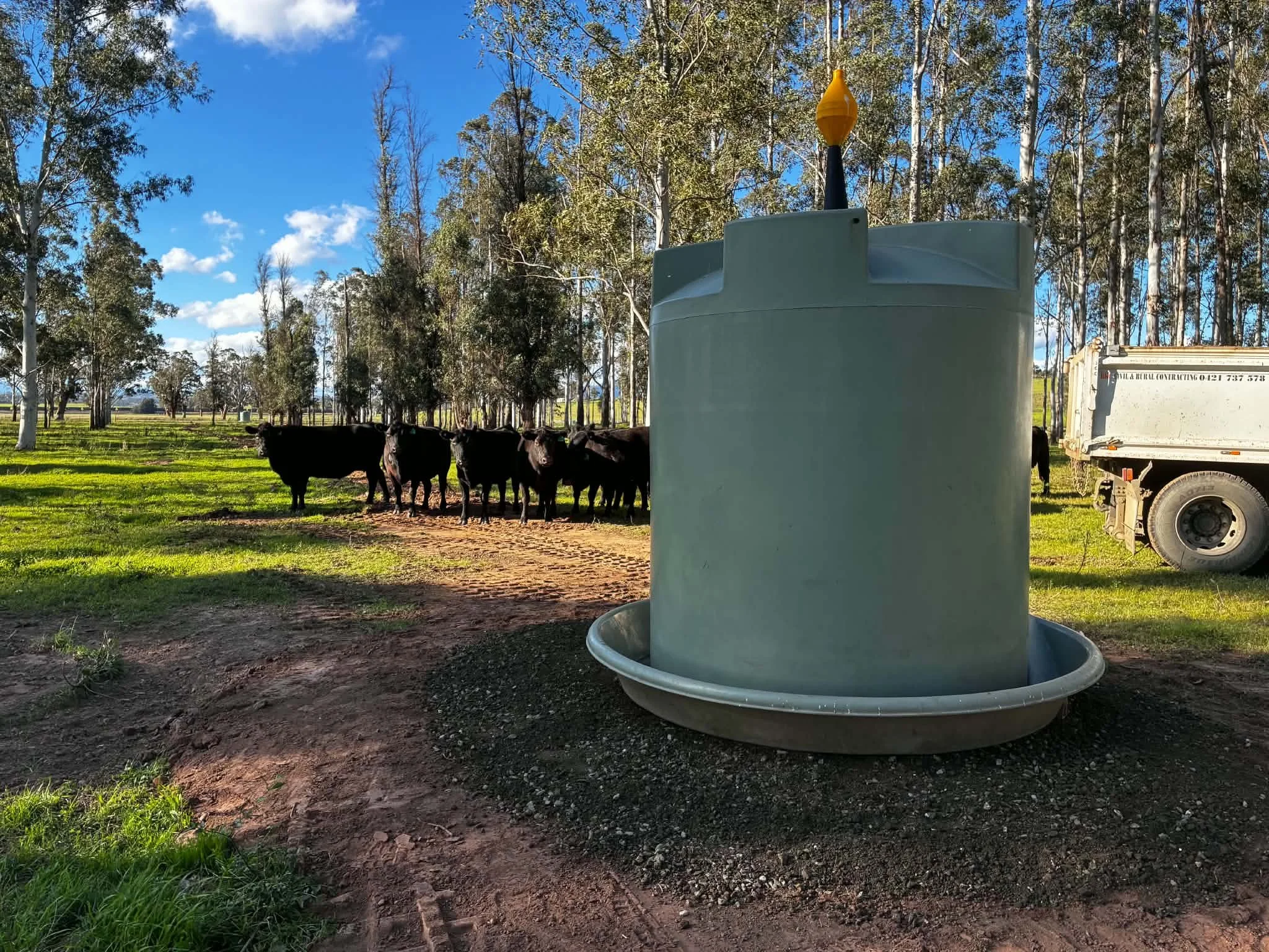 A large white and yellow cattle waterer with a drip tray at the bottom, in a grassy field with trees and a blue sky with some clouds in the background.