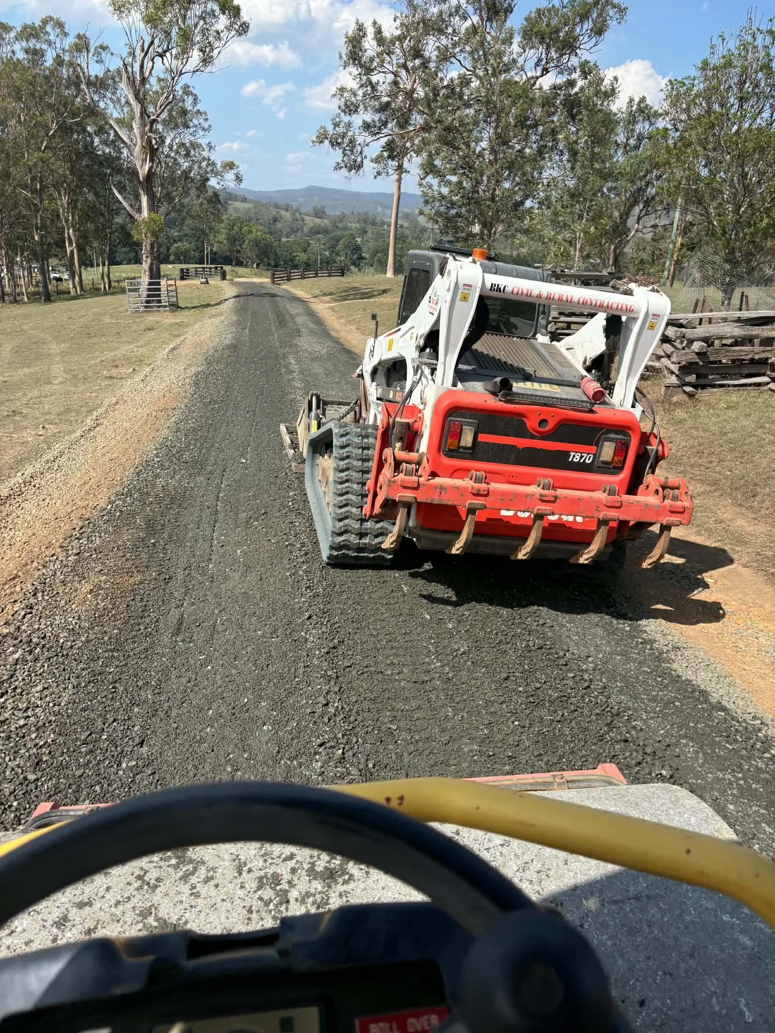 View from behind a vehicle with a black steering wheel on a rural dirt road, a red and white bulldozer in front, surrounded by trees and hills in the background, under a partly cloudy sky.