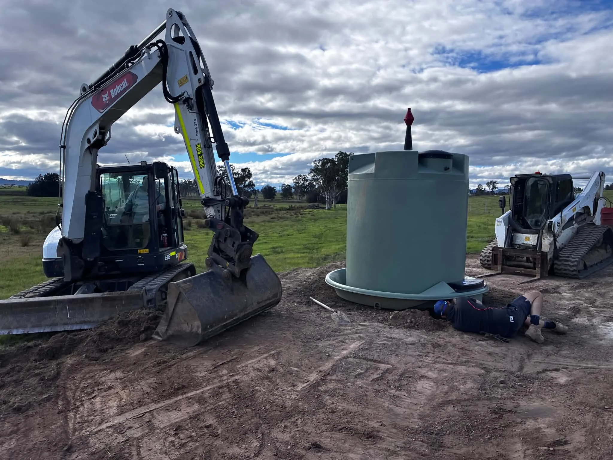 Construction site with two small excavators installing a large, gray, cylindrical structure with a red cone on top, in a grassy field under a cloudy sky.