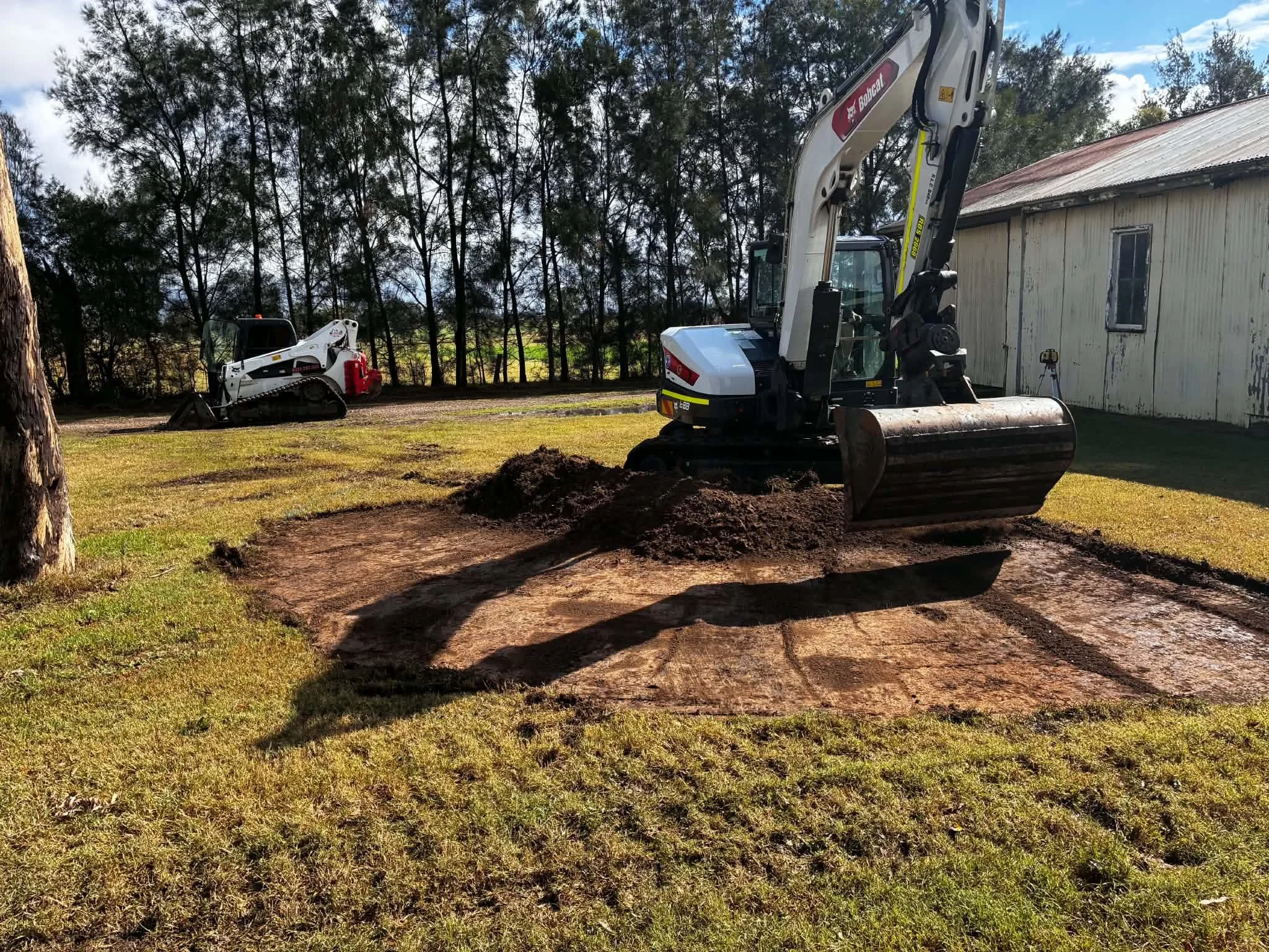 Construction site with a small excavator and a skid steer loader working on the grass near a white building and trees in the background.