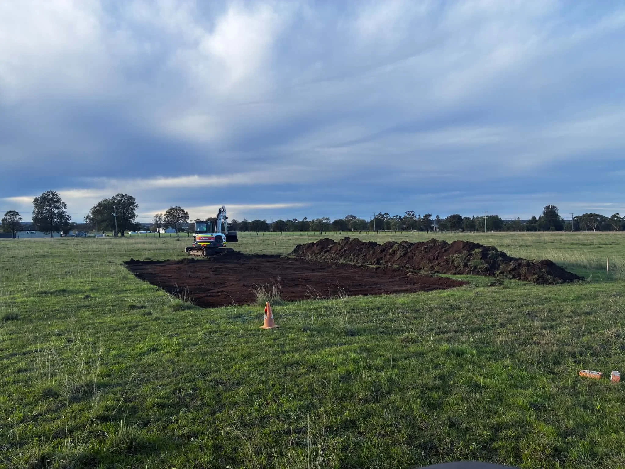 Construction site in a grassy field with a small excavator digging a trench, surrounded by dirt mounds and an orange traffic cone, under a cloudy sky.
