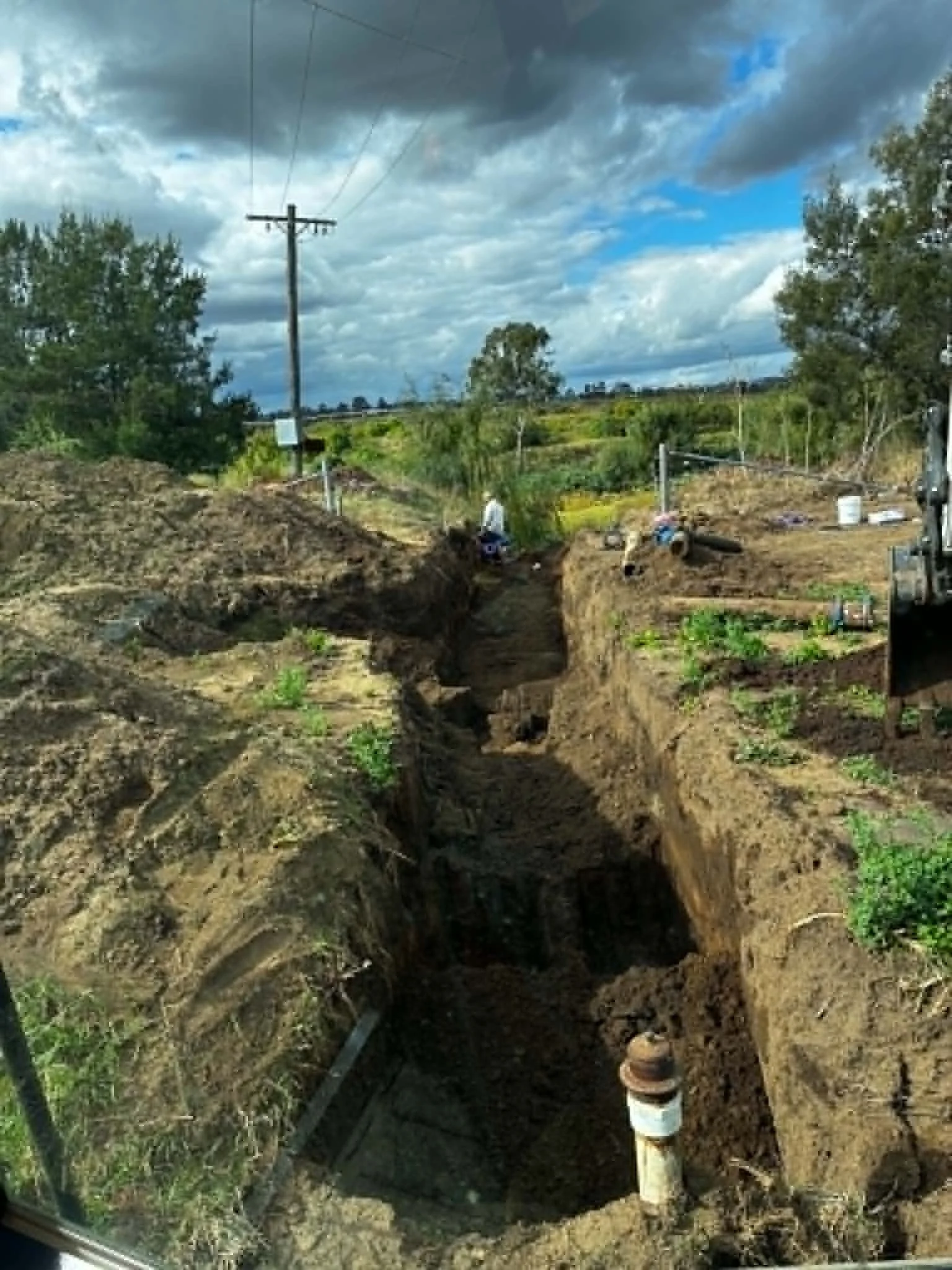 A deep trench dug in the ground with water pipes visible at the bottom, surrounded by soil and some green plants, with trees and a cloudy sky in the background.