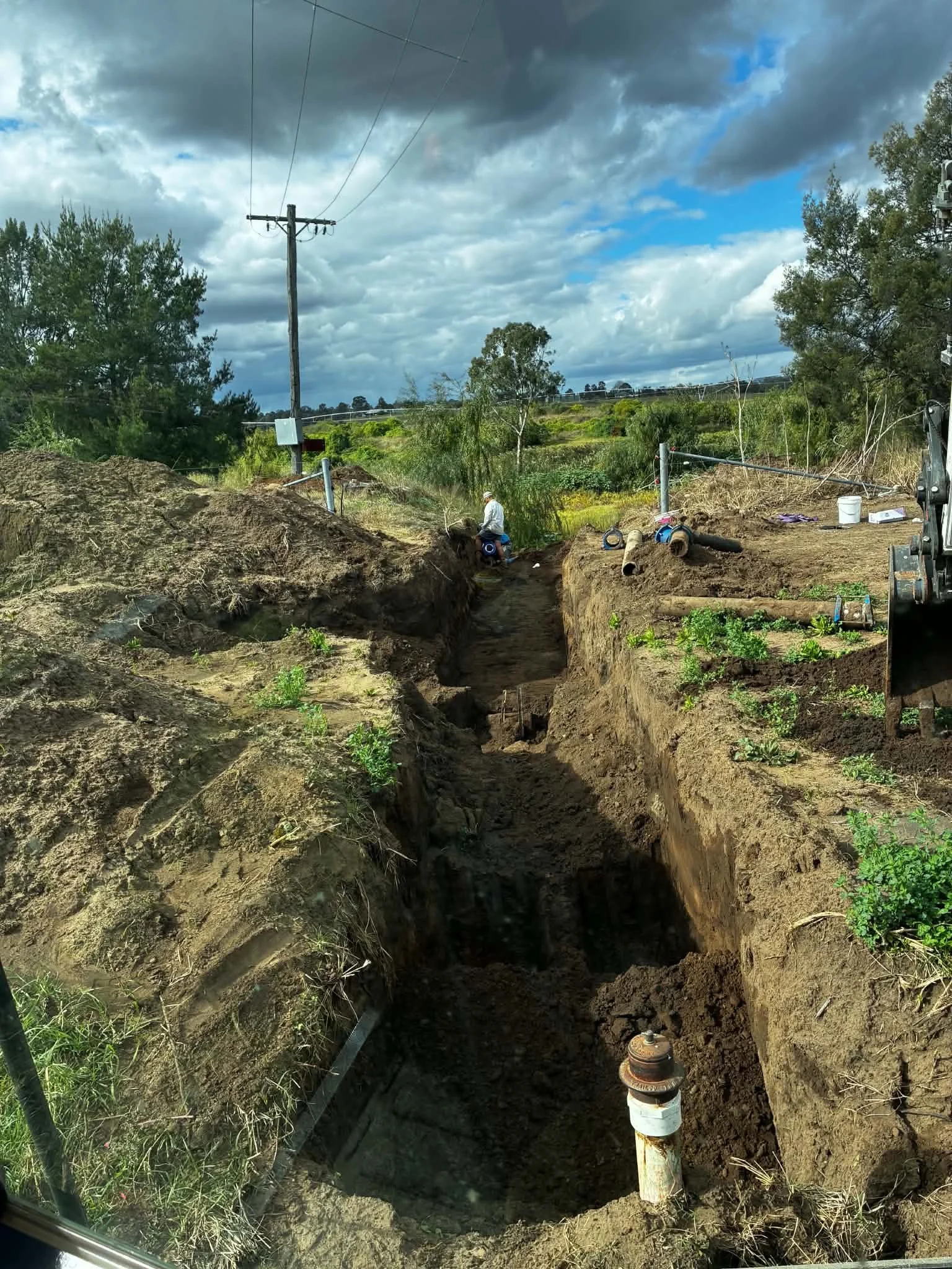 A person working in an open trench or excavation site with utility pipes and equipment, surrounded by green landscape and cloudy sky.