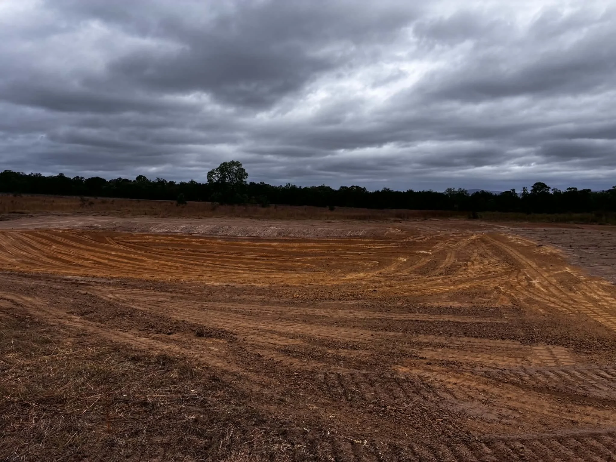A landscape scene with dark, cloudy sky and a flat, reddish-brown dirt field with visible tire tracks, bordered by a line of trees in the distance.