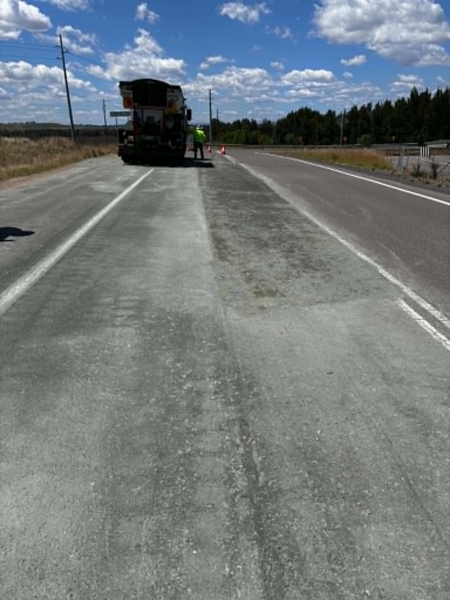 A road construction scene with a large truck and a worker in a neon vest. The truck is stopped on the side of a two-lane road under a bright blue sky with scattered clouds, and traffic cones are set up nearby.
