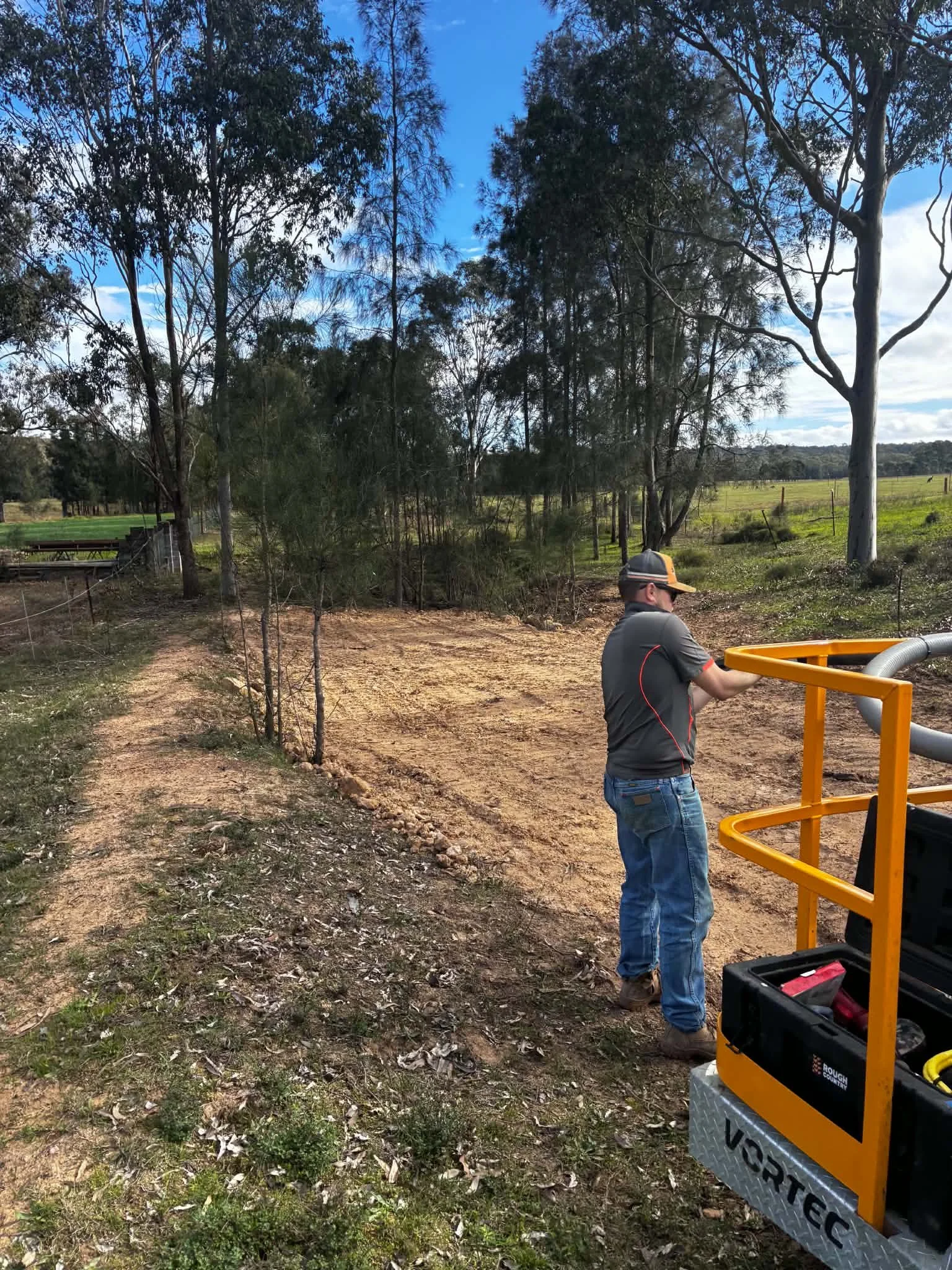 A man in a hat and gray shirt working outdoors with equipment, surrounded by trees and open field on a clear day.