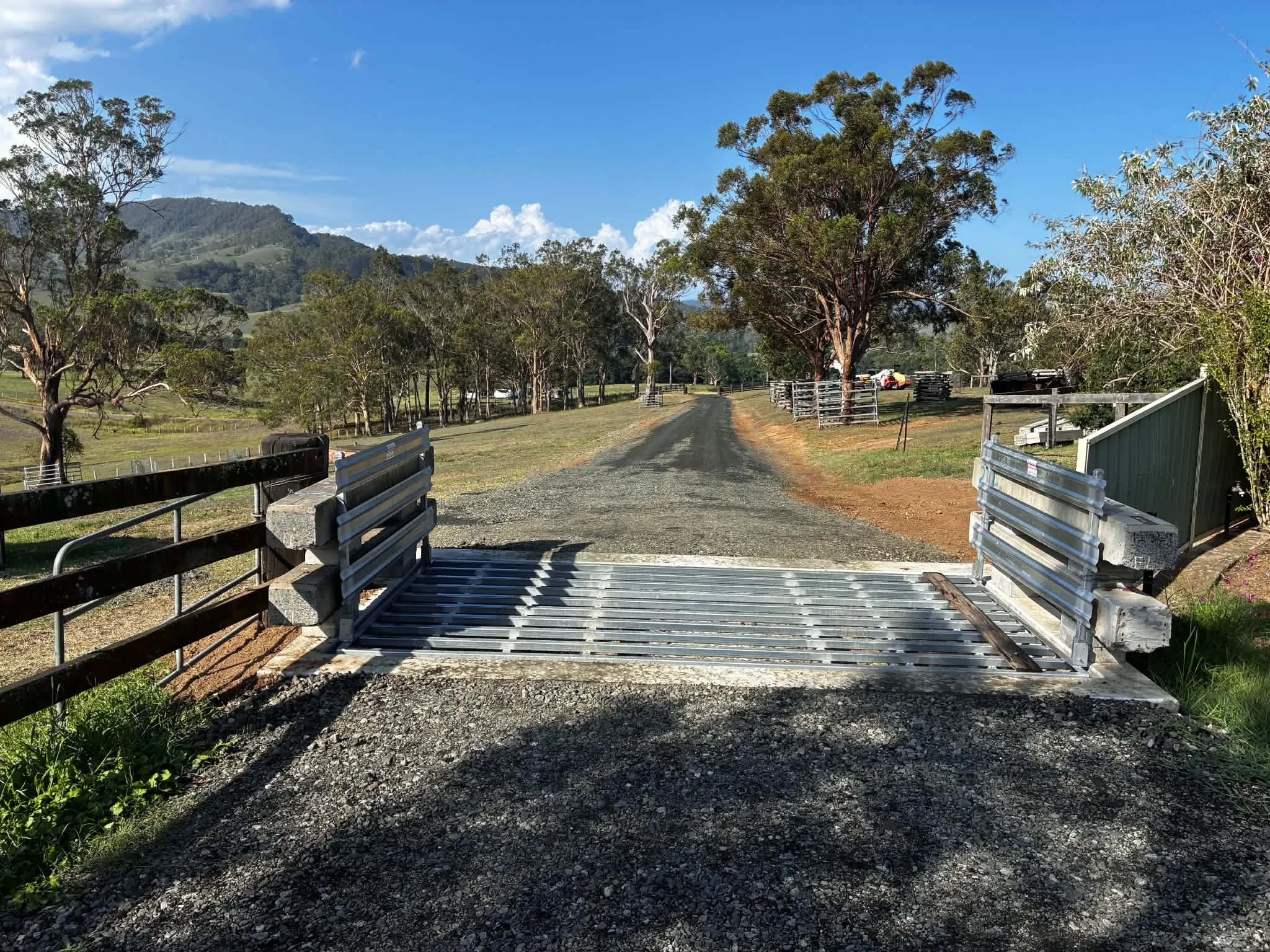 A rural scene with a gravel road, surrounded by trees and a mountain in the background, under a blue sky with white clouds.