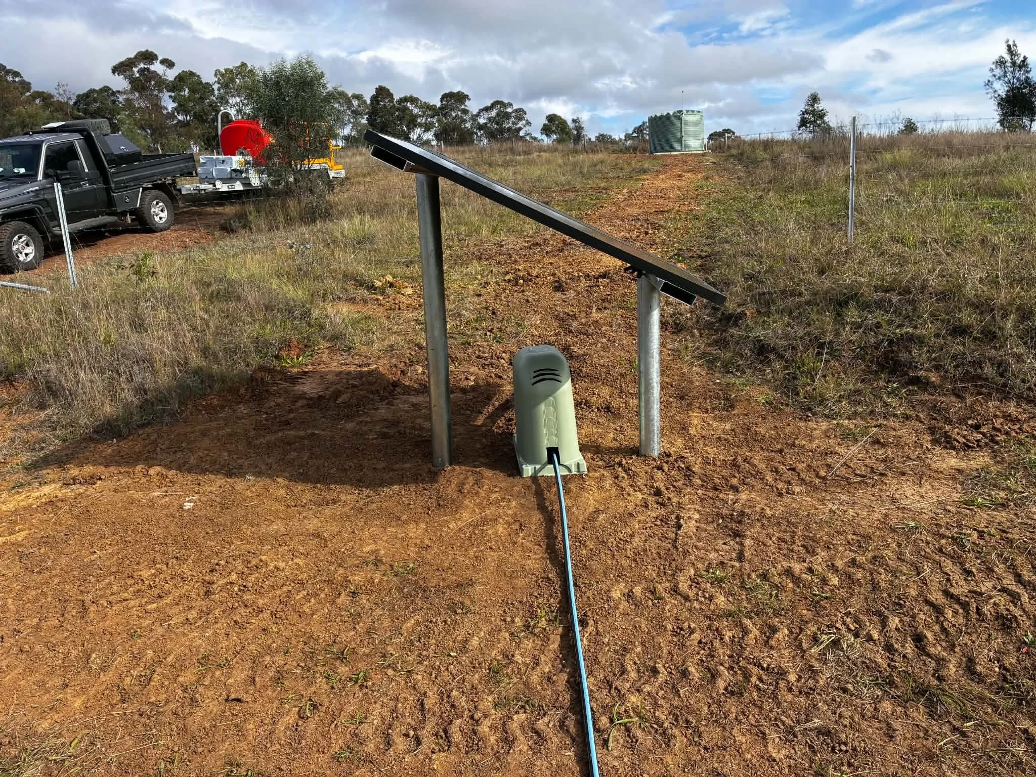 Solar panel in a dirt clearing with a blue cable connected to it, positioned in front of a fence, with trees and a water tank in the background.