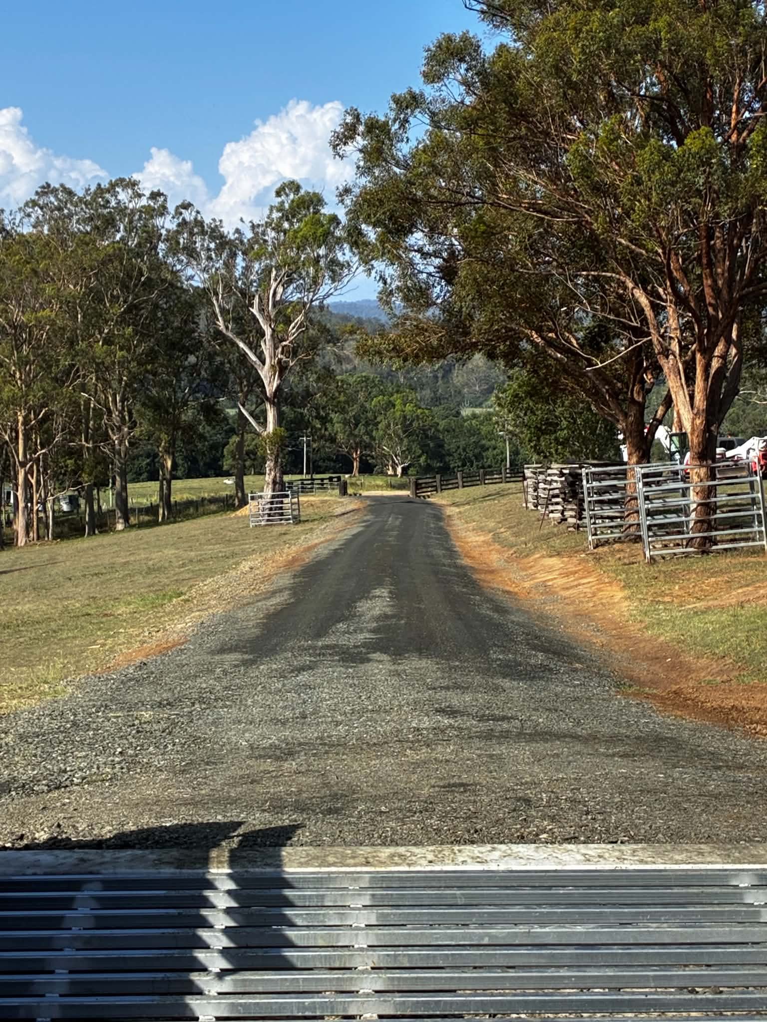 A rural road with a metal guardrail on the right side, lined with large trees and a grassy field, under a blue sky with scattered clouds.