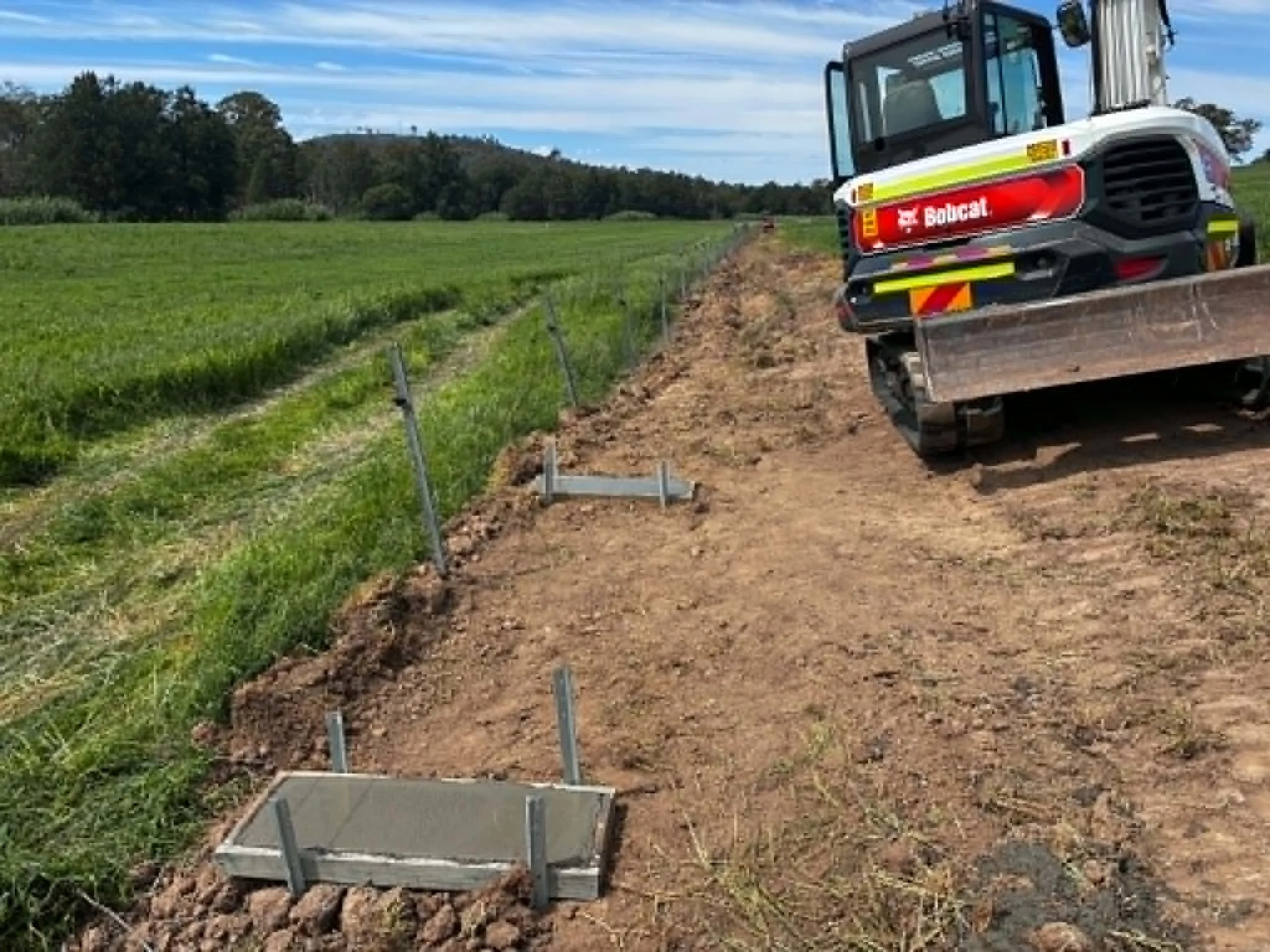 Construction site with a Bobcat skid-steer loader working on the ground next to a green field, with trees and a partly cloudy sky in the background.