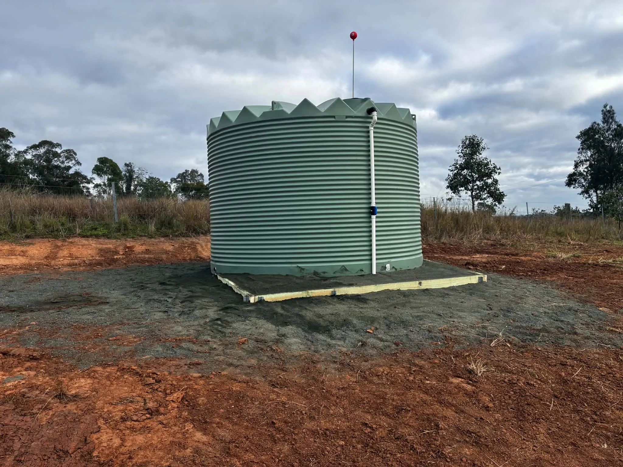 A large green water tank with a white pipe attached, situated outdoors on a concrete base, with trees and a cloudy sky in the background.