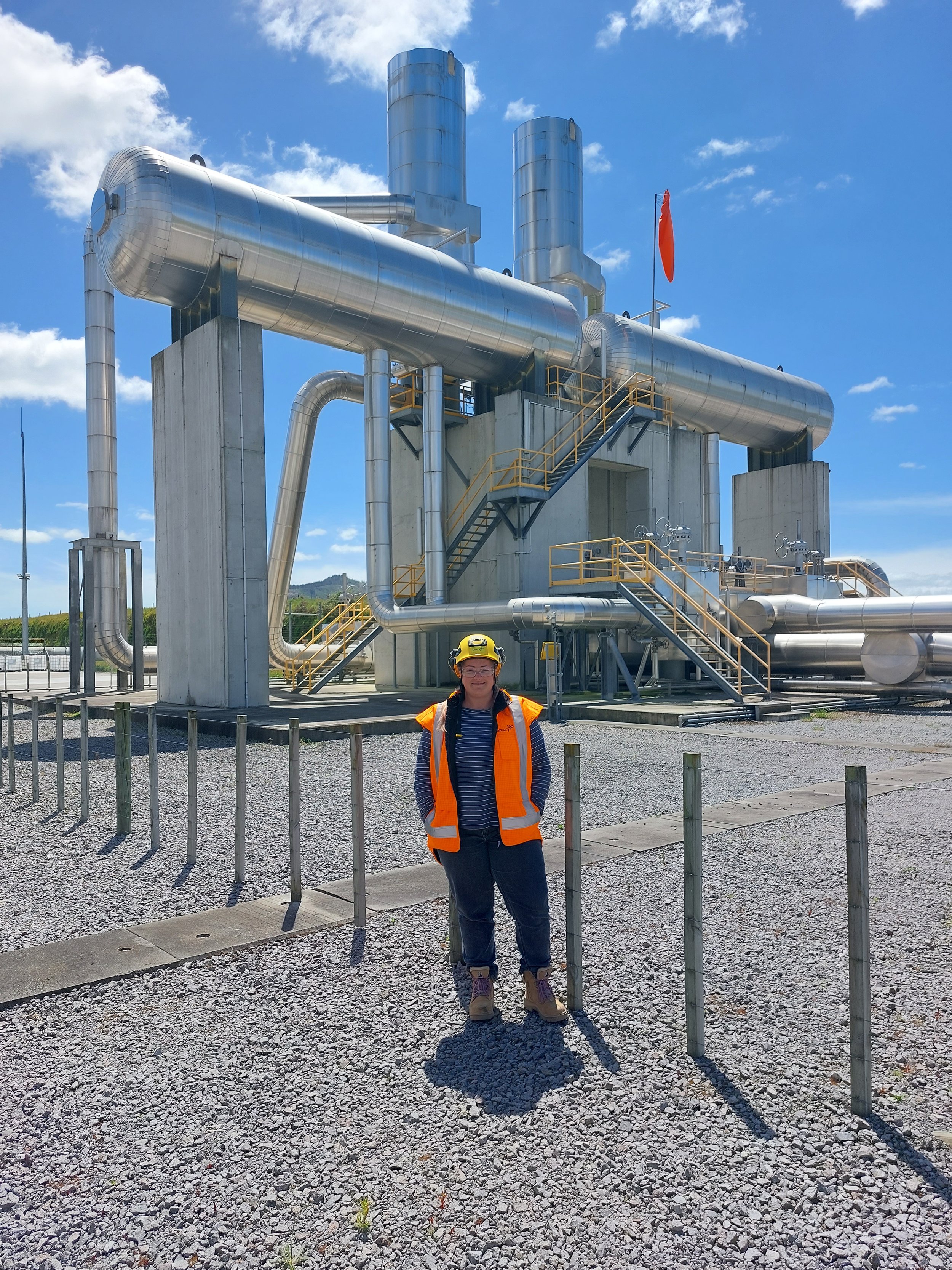 A woman wearing a yellow safety helmet and an orange safety vest standing outdoors in front of an industrial facility with large metal pipes and structures under a blue sky.