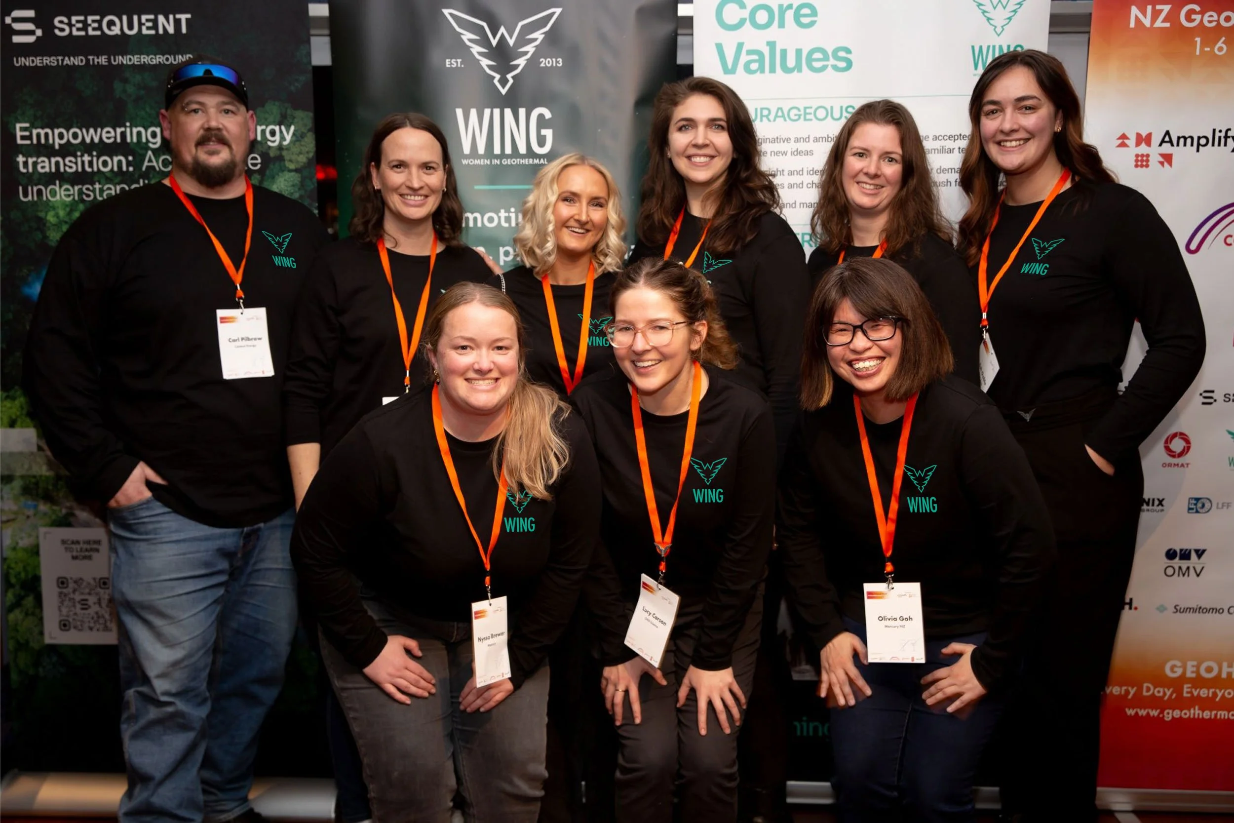 Group of ten smiling people at a conference, wearing black shirts with a teal wing logo and orange lanyards, standing in front of various banners with company logos and core values.