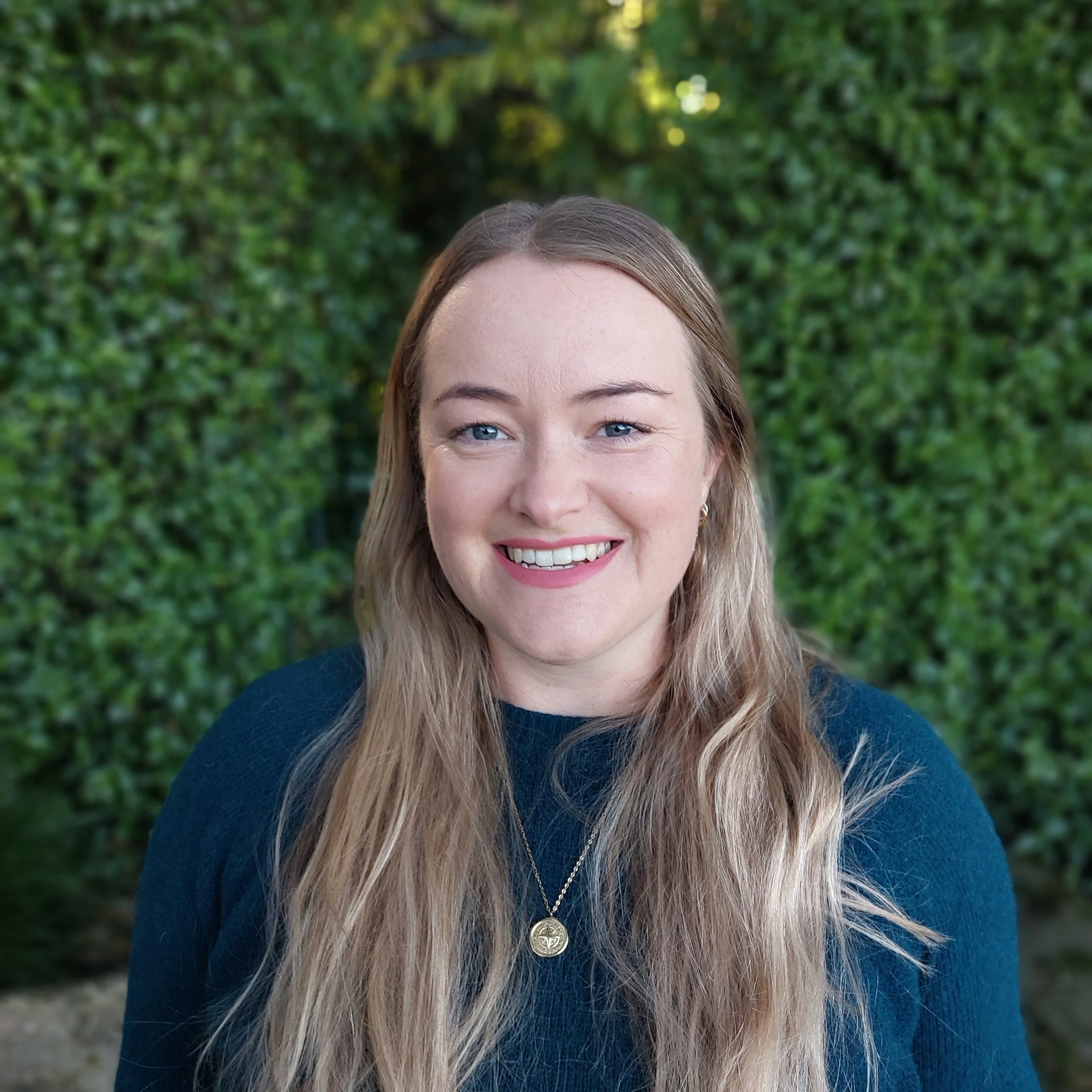 A smiling woman with long blonde hair wearing a dark blue top and a gold necklace, standing outdoors in front of a green foliage background.