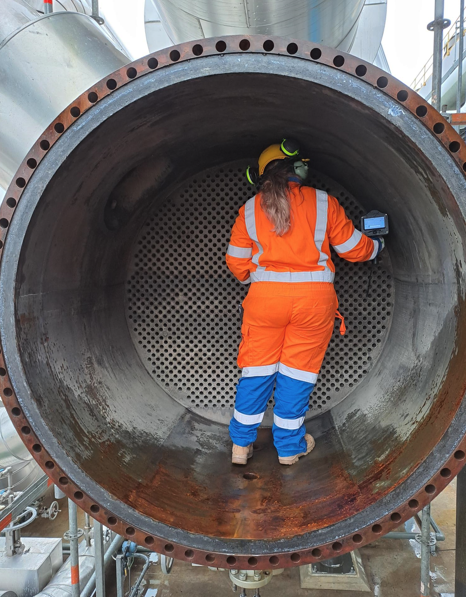 Person wearing orange safety gear and a yellow helmet inspecting the inside of a large metal pipe with a handheld device