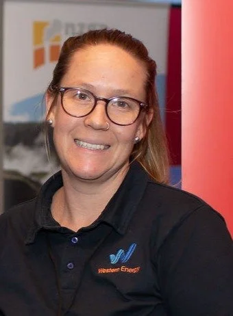 A woman with glasses and a friendly smile wearing a Western Energy polo shirt standing indoors.