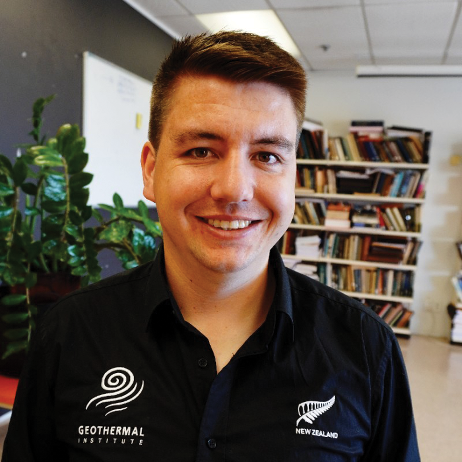 A young man in a black shirt with logos, smiling indoors with a bookshelf and a plant in the background.