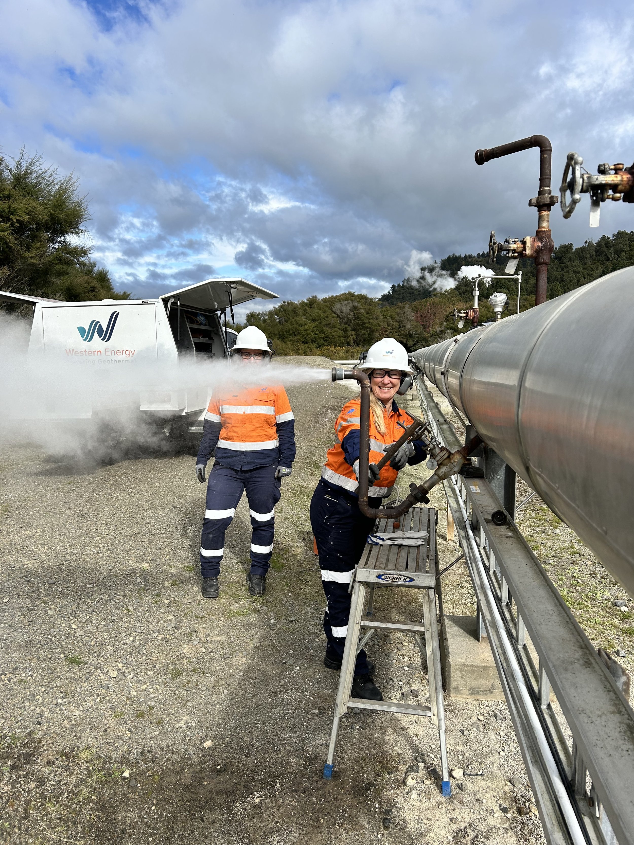 Two women working with industrial equipment outdoors, wearing hard hats and orange safety vests. One is smiling and operating a valve on a large metal pipe, while the other stands behind near a truck with the logo 'Western Energy'.