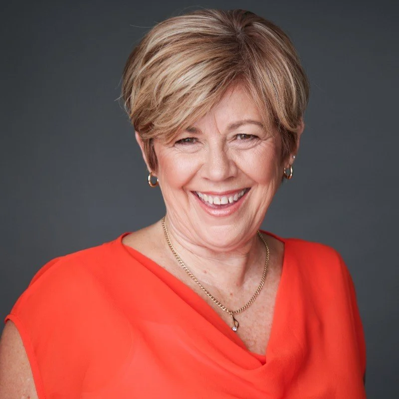 Smiling woman with short blonde hair, wearing a bright red top, gold necklace, and hoop earrings, against a dark gray background.
