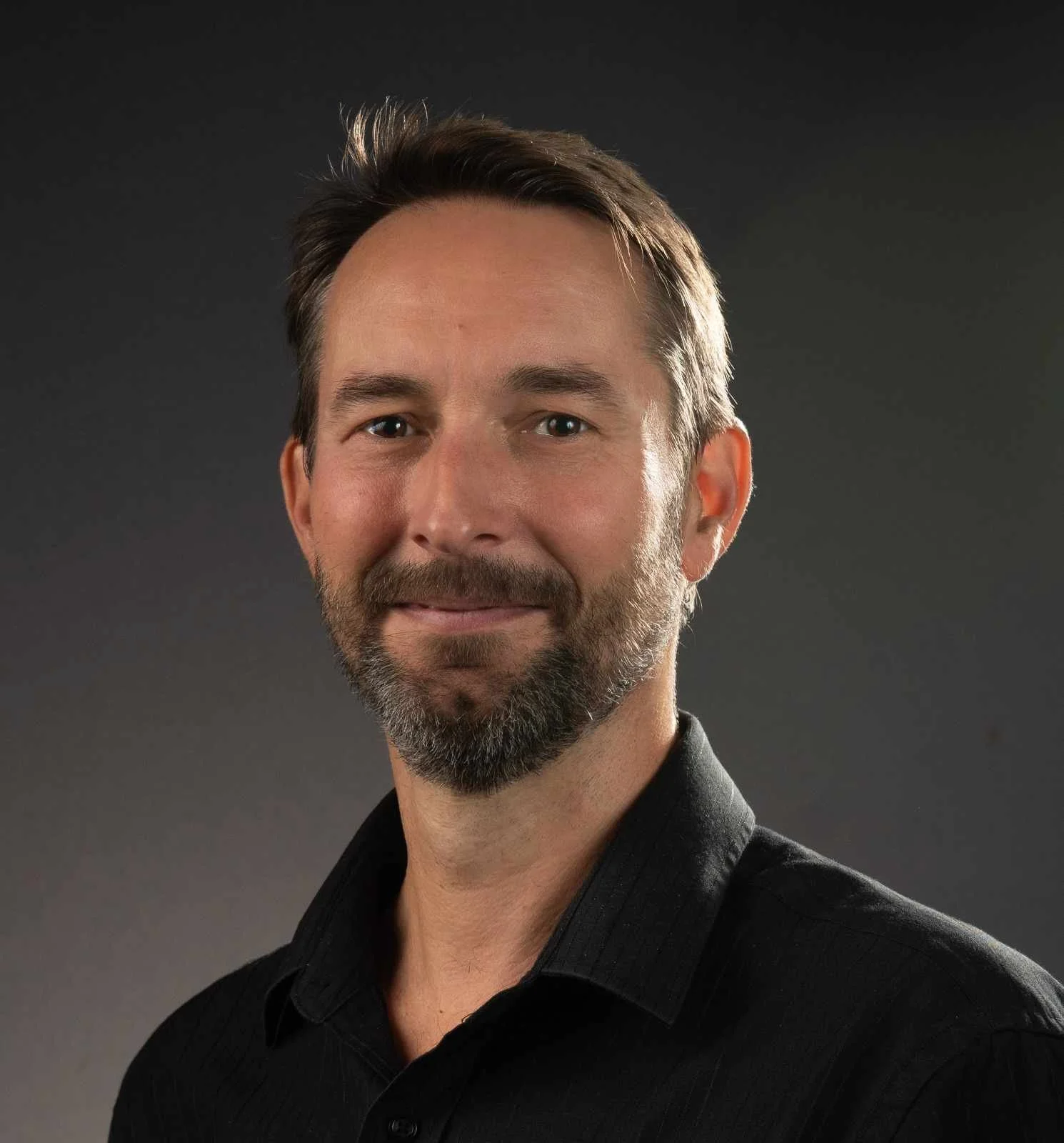 A middle-aged man with a beard and mustache, wearing a black shirt, smiling against a dark background.