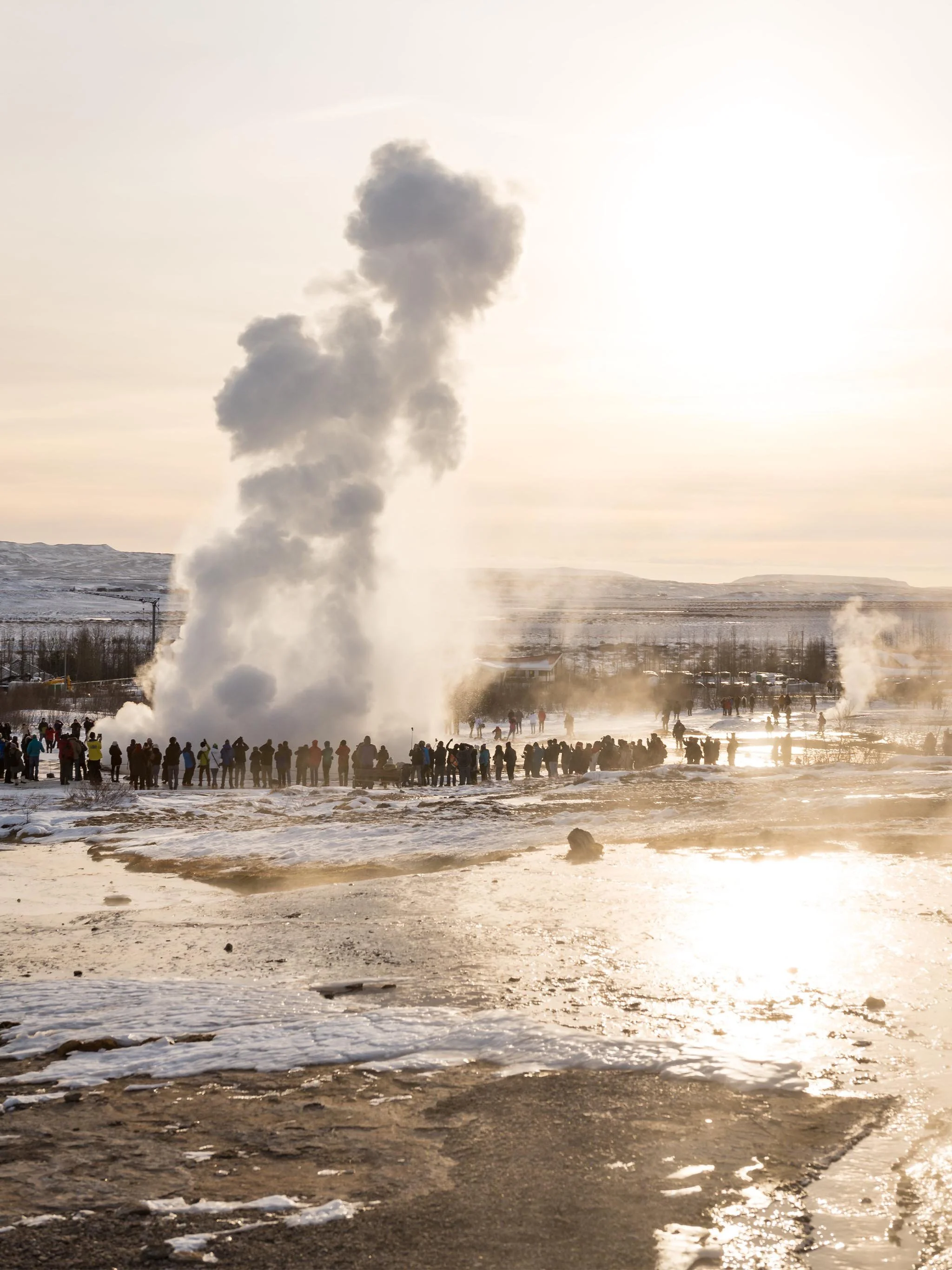 A crowd of people gathered around a geyser erupting in a snowy landscape during daylight. The geyser is releasing a tall plume of steam and water. Snow and ice cover the ground, with some patches of melted ground visible.