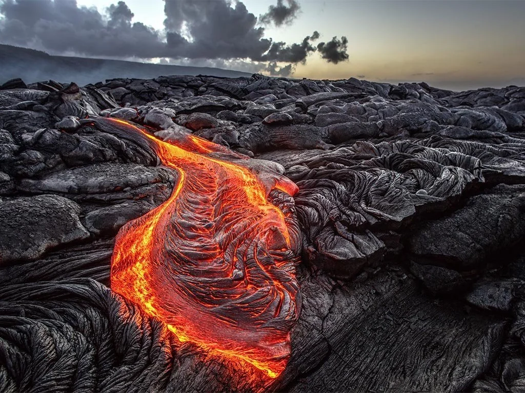 Flowing lava on black volcanic rocks during sunset, with dark clouds in the sky.
