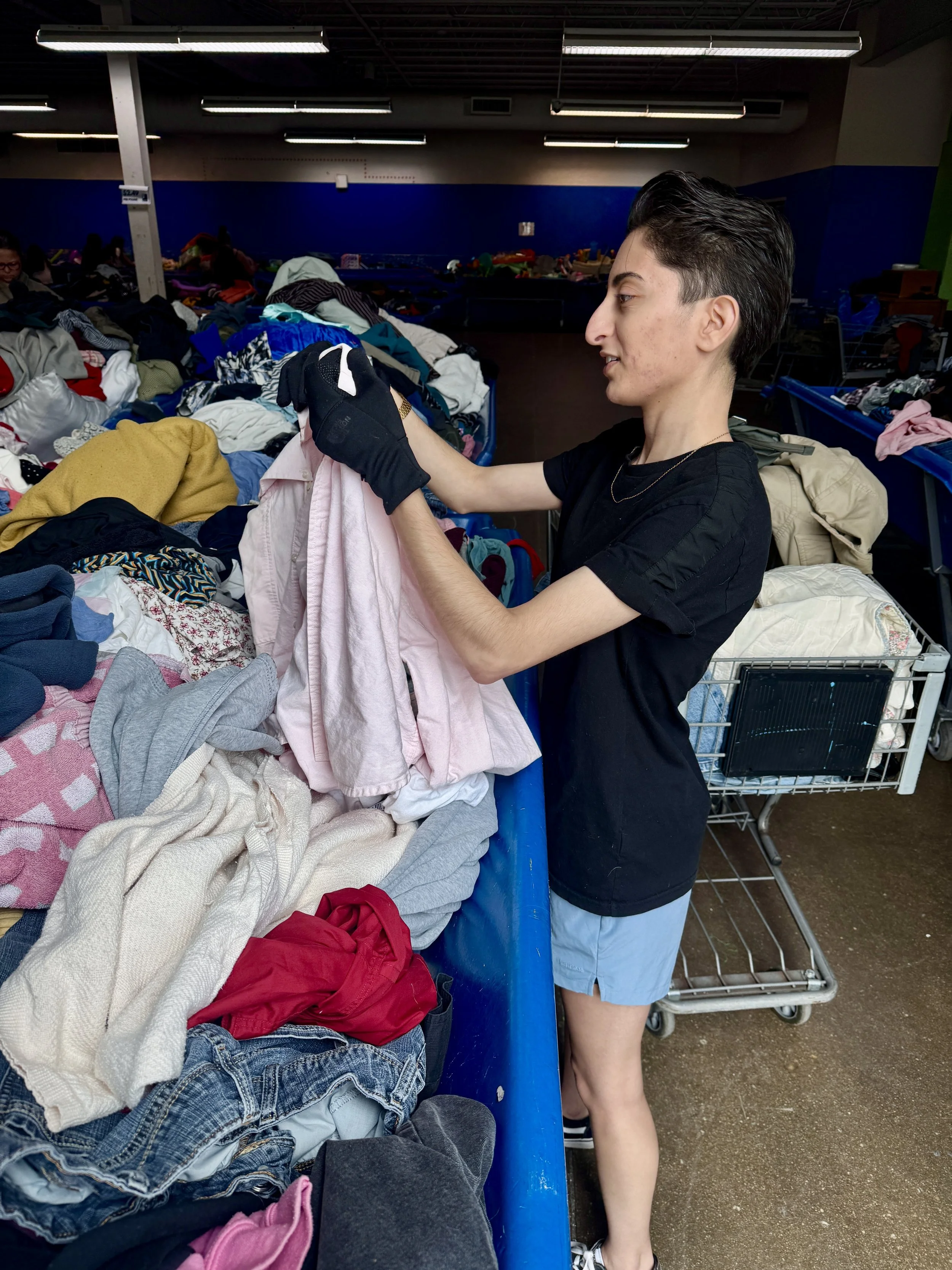 A young woman with short dark hair wearing a black t-shirt, light blue shorts, and black gloves sorting through clothes in a thrift store or donation center with tables filled with various secondhand clothing items.