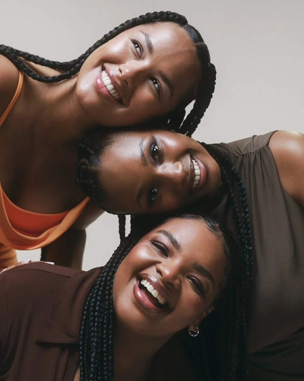 Three women have their heads stacked on top of one another. They're wearing braids and smiling.