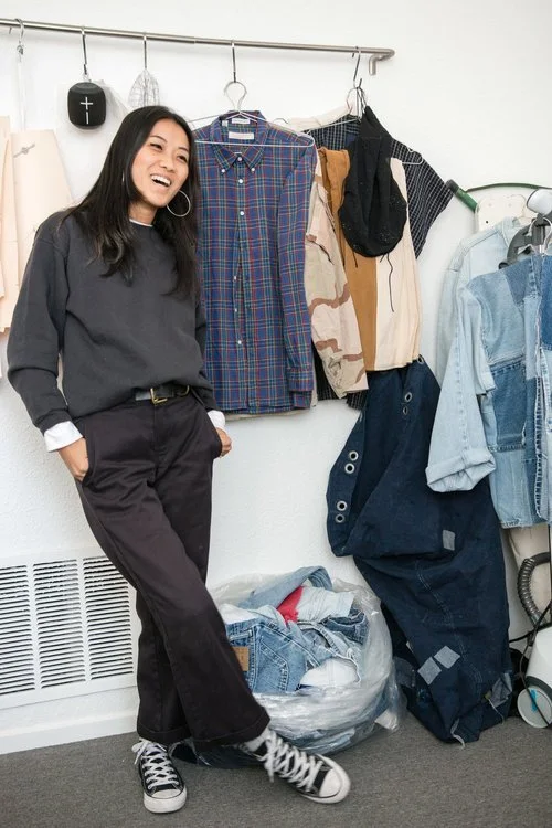 Natasha Halesworth of The Consistency Project smiling in front of recycled clothing. Wearing black pants, black chuck taylors, and a grey sweatshirt.